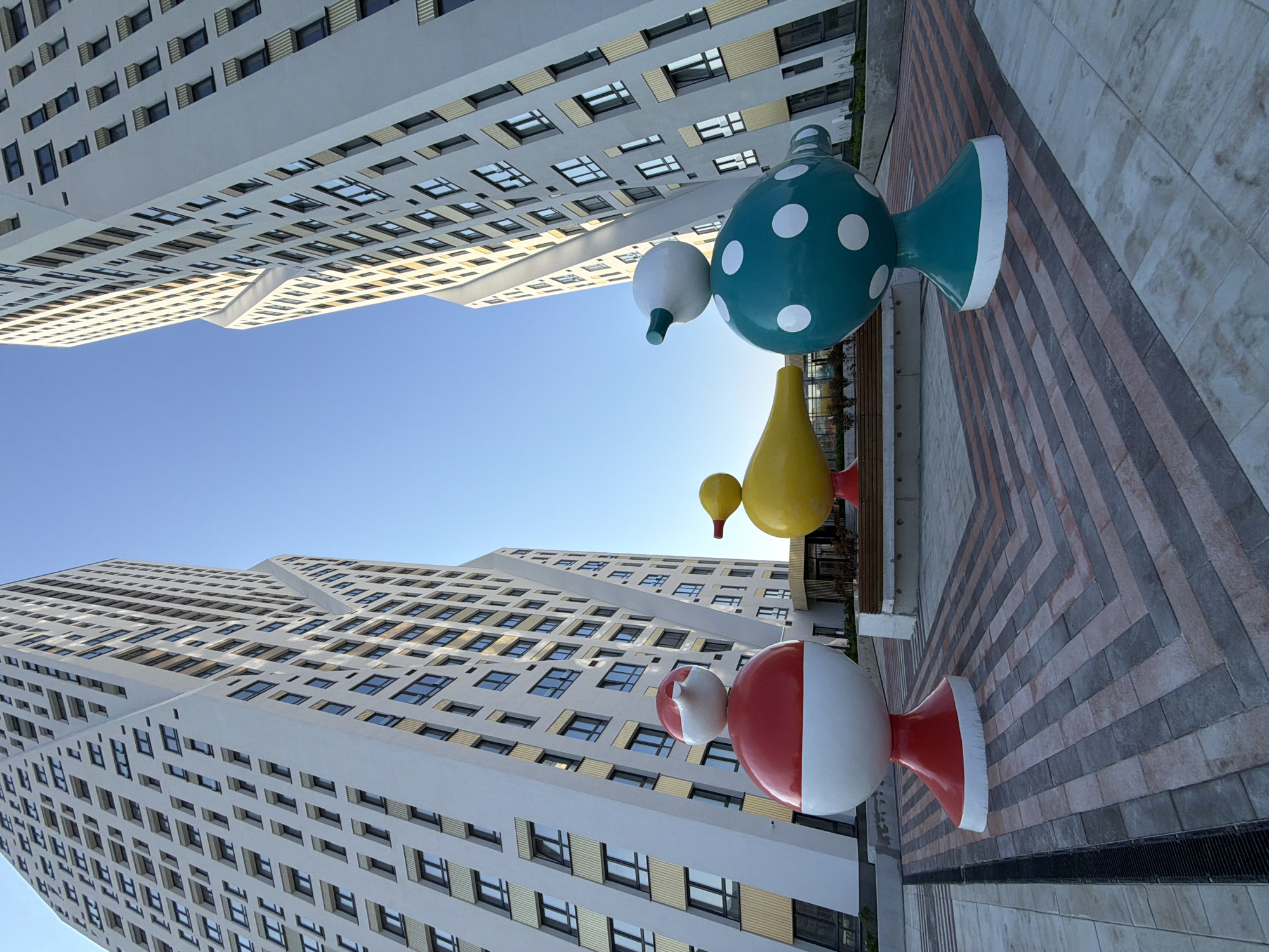 A low-angle, wide shot captures a public plaza in Yekaterinburg, Russia, featuring a vibrant outdoor art installation against the backdrop of two imposing modern apartment buildings. The plaza's ground is paved with a geometric pattern of rectangular stones in shades of gray, pink, and beige, with a dark grate running along the left edge. In the foreground and mid-ground stand three whimsical, large-scale sculptures that resemble stylized birds.
The leftmost sculpture is predominantly red with a white band and a smaller white spherical head, evoking a sense of a festive character. Next to it is a bright yellow, duck-like sculpture with a red beak. To the right is a teal sculpture adorned with white polka dots, also featuring a white spherical head and beak-like protrusion.
The two apartment buildings, designed in a contemporary architectural style with alternating bands of light-colored facade and windows, flank the plaza, creating a sense of enclosure. Their facades have a striking, angular design that twists as they ascend. The sky above is a clear, bright blue. No people are visible in the scene, and the overall impression is one of modern urban design combined with playful public art. The location is identified as the area around the UGTU (Ural State Technical University) campus, specifically near the new residential complex "Nekrasov," in Yekaterinburg, Russia.