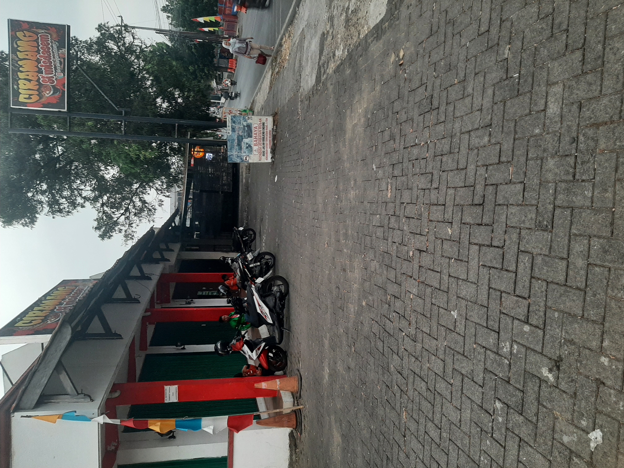 The image depicts a street scene in front of a building, identified as "CIRAWANG SI MIEDASEM," in Purwanegara, Indonesia. The building has red pillars and green metal shutters, with colorful flags hanging on the left side. Several motorcycles are parked on the brick-paved sidewalk in front of the building, with a few individuals appearing to be seated or standing near them. A tall sign with the building's name is visible on a pole. Further down the street, a woman in a hijab is walking on the sidewalk, carrying a red bag. A sign on the sidewalk indicates a road hazard. The environment appears to be an urban or semi-urban setting with trees and utility poles present. The sky is overcast, suggesting a cloudy day.