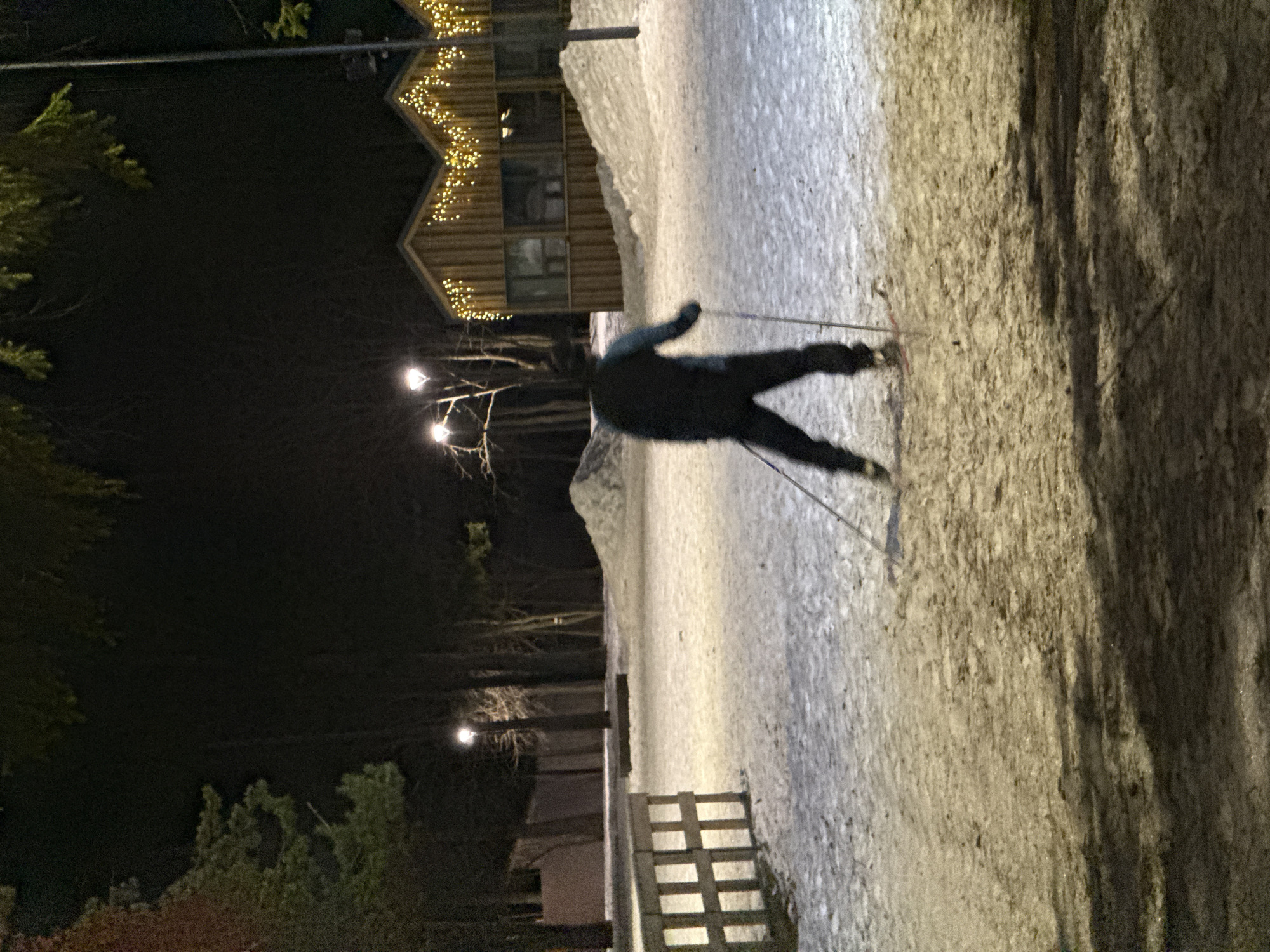 A cross-country skier glides across a snow-covered track at night. The skier, clad in dark clothing, is silhouetted against the illuminated path. The scene includes a building decorated with lights, trees, lampposts, and a wooden fence. The location is likely a park or recreational area in Votkinsk, Russia, known for its winter activities.