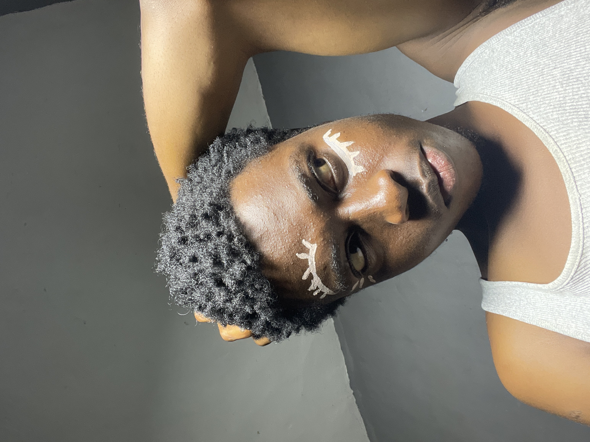 A person with dark skin and short, curly hair is looking towards the camera. They are wearing a white tank top and have white paint applied in a decorative pattern around their eyes and on their forehead. The person's right arm is raised and bent, with their hand resting on the back of their head. The background is a plain, gray wall. This scene is located in Nkpor, Nigeria.