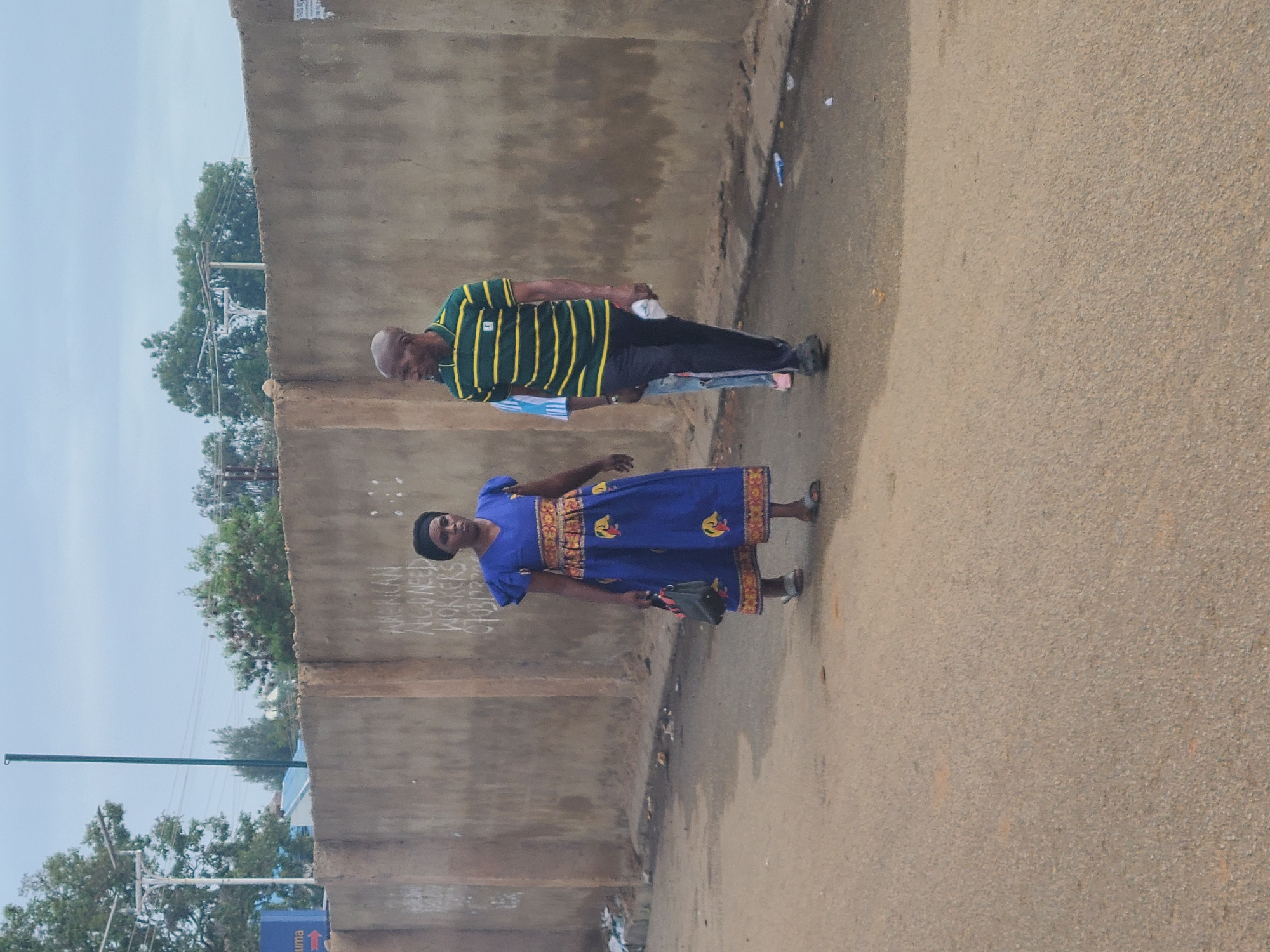 Two individuals are walking on a paved road next to a concrete wall in Jos, Nigeria. The wall has graffiti reading "AMERICAN NURSES/WORKERS NEED AFRICA." One individual is a man wearing a green and yellow striped shirt and black pants. The other is a woman wearing a blue dress with patterned trim. The environment includes trees, a cloudy sky, and utility poles.