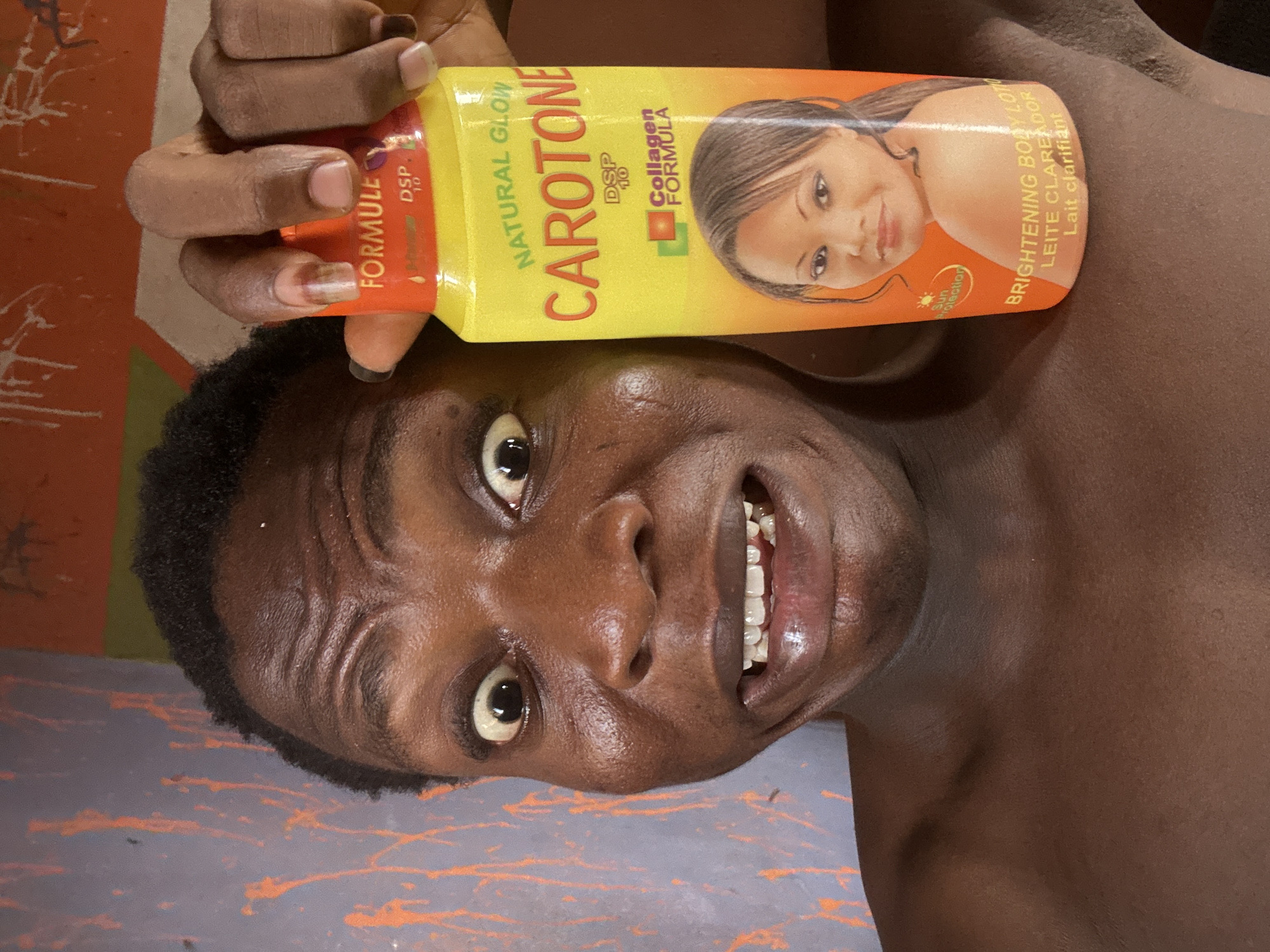 A shirtless person with dark skin and short, curly hair is lying on their back, holding a bottle of "CAROTONE" brightening body lotion. The bottle is yellow and orange and features an image of a light-skinned woman on the label. The person is smiling and looking directly at the camera with wide eyes. The background is a surface with orange and gray paint splatter.