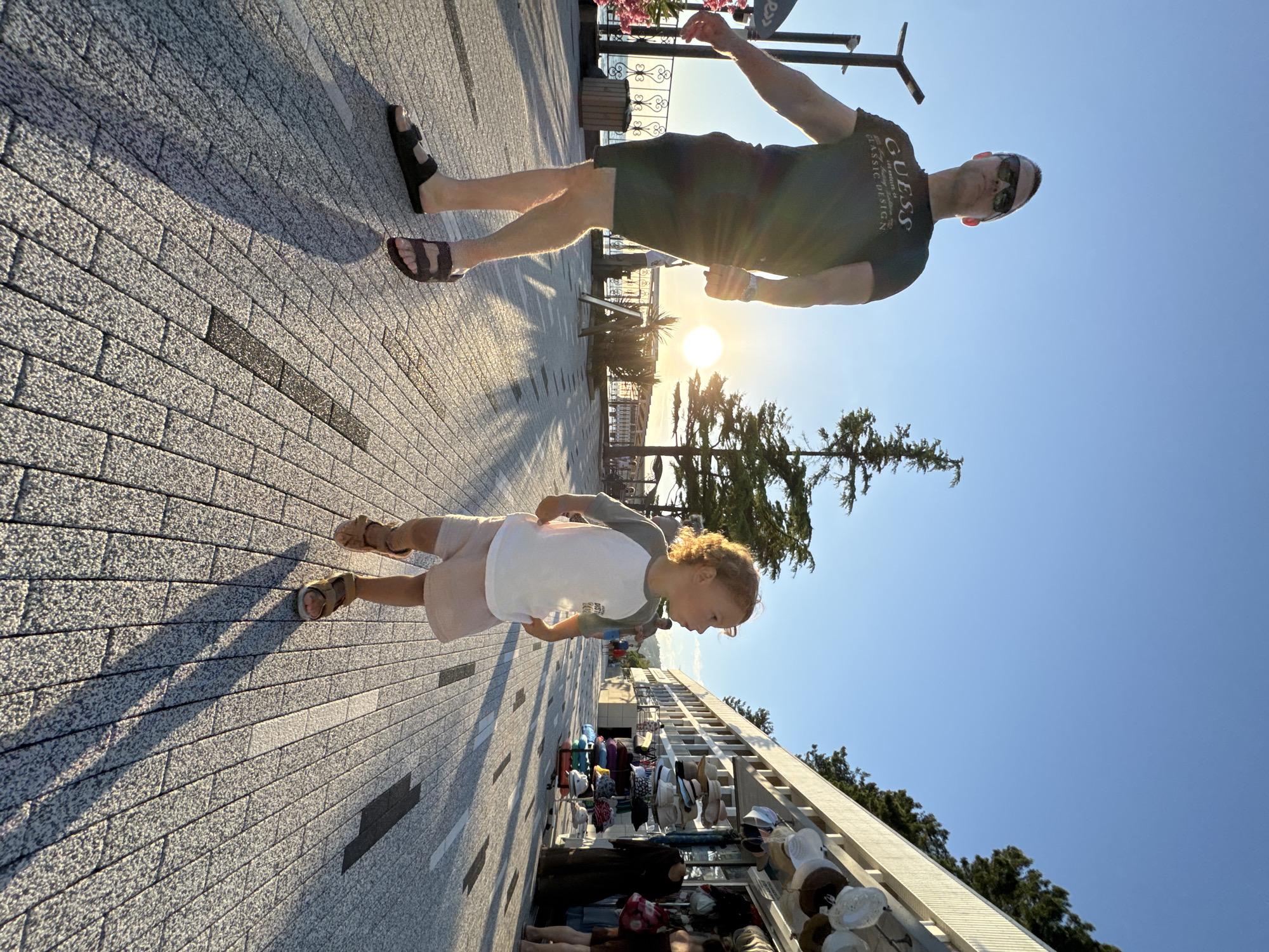 In the city of Sochi, Russia, on a bright, sunny day, a man and a young child are walking along a paved promenade. The man, wearing a black t-shirt, dark shorts, and black sandals, is in the foreground on the left, facing forward and slightly to the right, with a slight smile on his face. He is wearing sunglasses and his hair is short. The child, in the mid-ground on the right, is facing to the right and looking ahead with a curious expression. The child is wearing a white and gray t-shirt with a graphic print, light pink shorts, and brown sandals.
The promenade is paved with rectangular gray bricks and has black and white markings. On the left side of the promenade, there is a railing overlooking the sea, and a trash can. Further back, a tall, thin tree stands against the clear blue sky. The sun is low in the sky, casting long shadows across the promenade.
On the right side of the promenade, there are shops with awnings, displaying hats and other merchandise. Several people are visible in the background, some browsing the shops and others walking along the promenade. The architecture of the buildings behind the shops suggests a resort or tourist area. The overall atmosphere is one of leisure and a pleasant evening stroll.