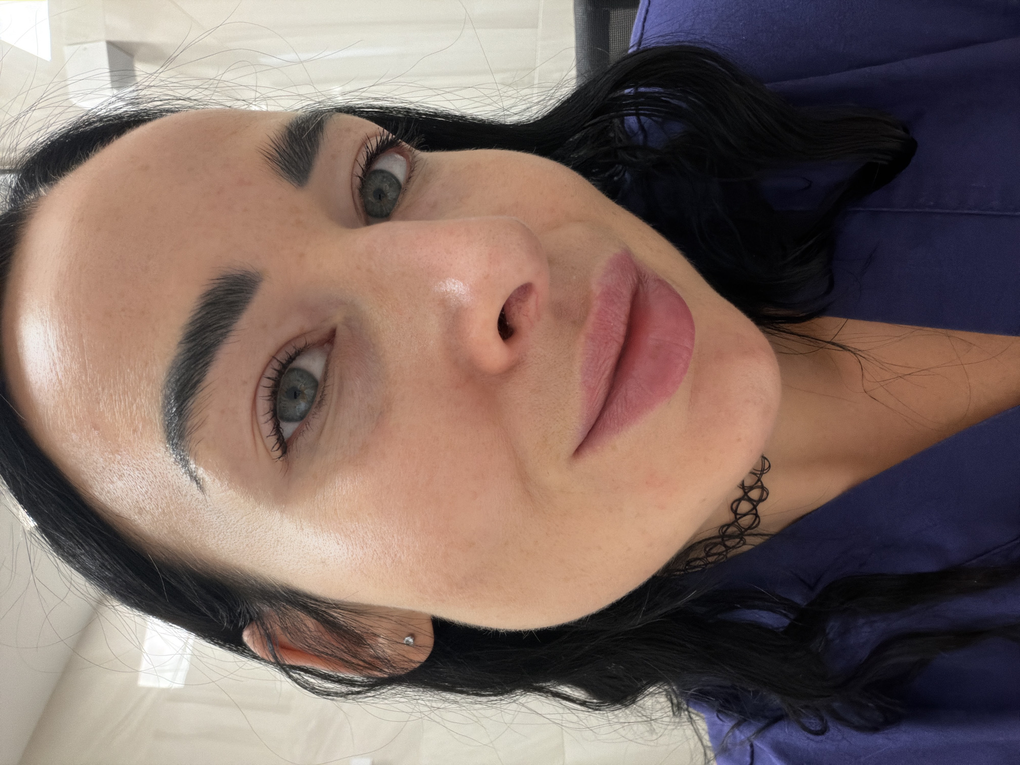 A close-up, rotated view of a woman's face. She has dark hair, light skin, defined eyebrows, and light-colored eyes. She is wearing a dark-colored garment. A delicate choker-style necklace is visible on her neck. The background appears to be a neutral-colored ceiling and a dark fabric. Location information is not present.