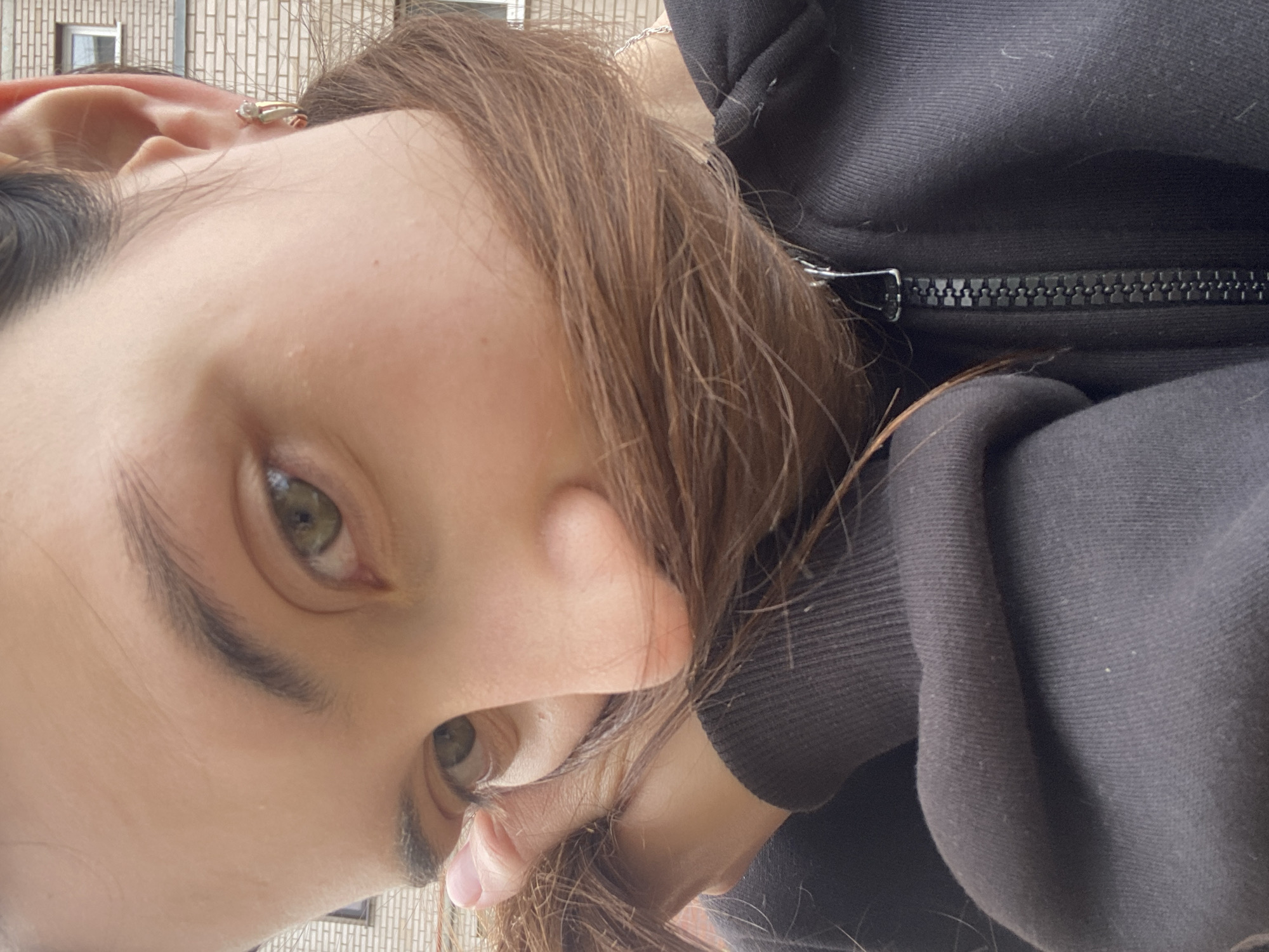The image is a close-up, outdoor selfie of a young woman with brown hair and green eyes. She is wearing a black hoodie, and her hair is obscuring the lower half of her face. The background appears to be an urban environment, with a brick building and a window visible. The lighting suggests it is daytime. The woman's right hand is visible, holding some of her hair. She is wearing a small hoop earring in her right ear. The image was taken in Votkinsk, Russia.