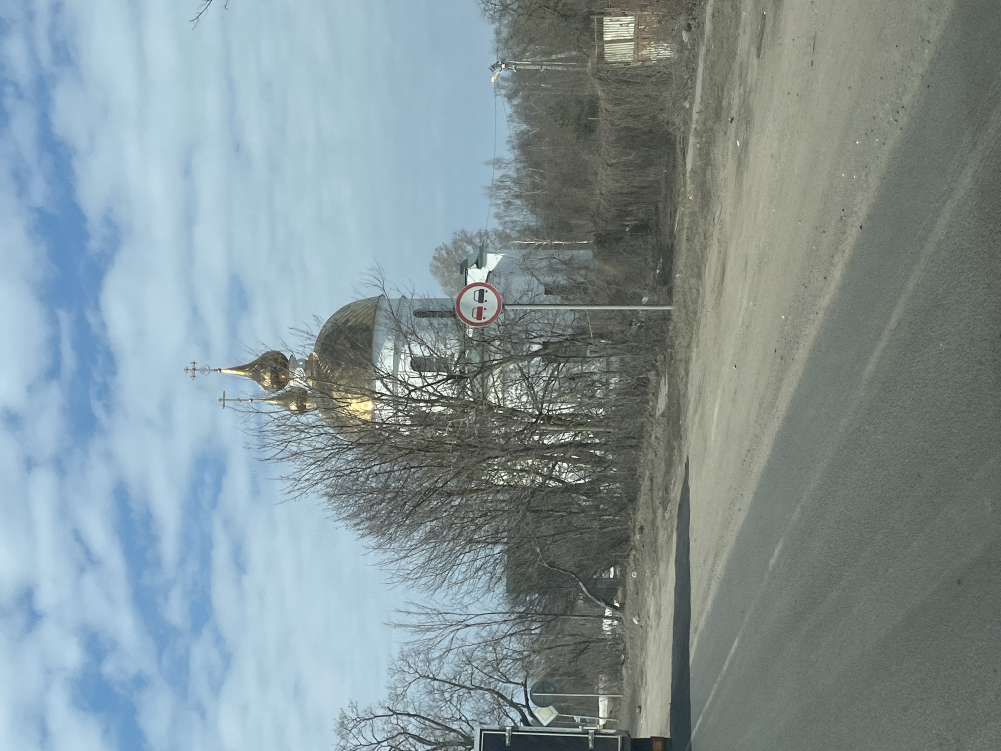The image displays an outdoor scene in what appears to be late winter or early spring, with a prominent Russian Orthodox church as the central background element. The location is stated as Pokrovskoe, Russia.
In the foreground, an asphalt road occupies the lower left quadrant, displaying a faint white dashed centerline. To its right, a wide, unpaved dirt or gravel shoulder extends, indicating a roadside environment.
The main subject is a white church situated in the mid-ground, featuring at least two onion-shaped golden domes topped with Orthodox crosses. The church's white facade is partially obscured by numerous deciduous trees and dense brush, all bare of leaves.
To the right of the asphalt road, positioned between the road and the church, a circular regulatory road sign is visible on a grey pole. The sign displays a red circle with a diagonal line, depicting two cars (a red one on the left, a black one on the right), signifying "No Overtaking" or "No Passing."
Further to the right, utility poles with power lines are present. A rustic fence-like structure, heavily overgrown with dry vines or brush, defines a boundary in the right background. The sky is partially cloudy, with patches of blue visible amidst white, scattered clouds. No individuals or active interactions are discernible in the scene.