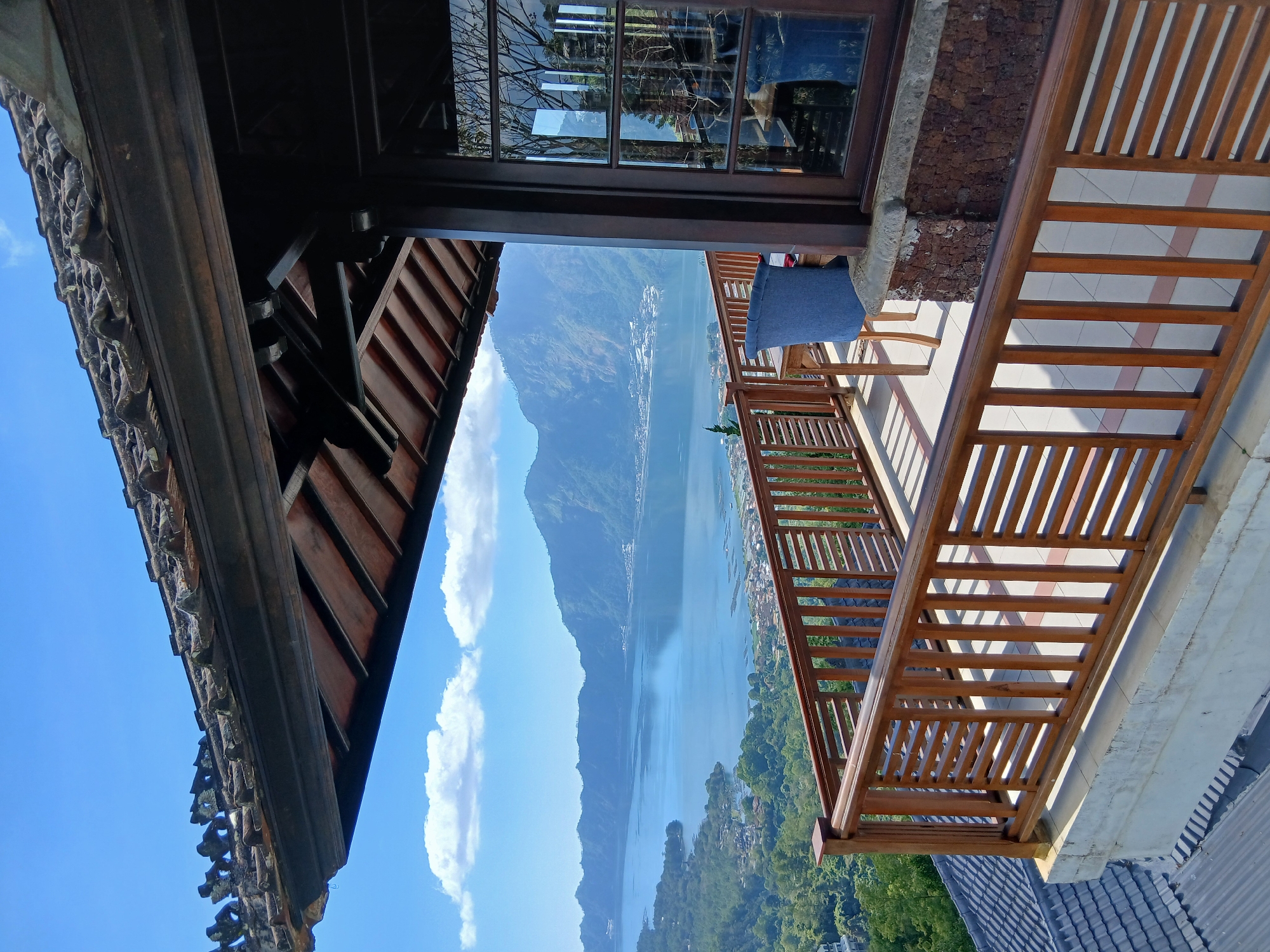 The image depicts a scenic view from a wooden balcony overlooking a large lake with mountains in the background, likely Lake Batur in Kintamani, Indonesia. The foreground features a traditional Balinese wooden railing and a portion of a building's roof with clay tiles. Beyond the railing, a table and a blue upholstered chair are visible on the tiled balcony floor. The lake, a prominent feature, is surrounded by lush green hills and dotted with small villages and fish farms. Majestic mountains form a dramatic backdrop to the tranquil waters, under a clear blue sky with scattered white clouds. The scene is bathed in bright sunlight, casting shadows on the balcony. No people are present in the image.