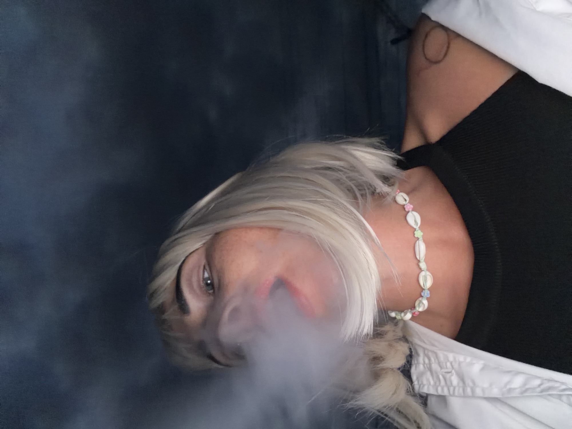 A low-angle, close-up shot shows a person with blonde hair and fair skin exhaling a cloud of white vapor. The person is wearing a black tank top, a white open shirt, and a necklace made of shells. The background is dark and out of focus, suggesting an indoor or nighttime setting. There is a faint circular tattoo visible on the person's left shoulder. The image does not contain any discernible features indicating a specific location in Sochi, Russia.
