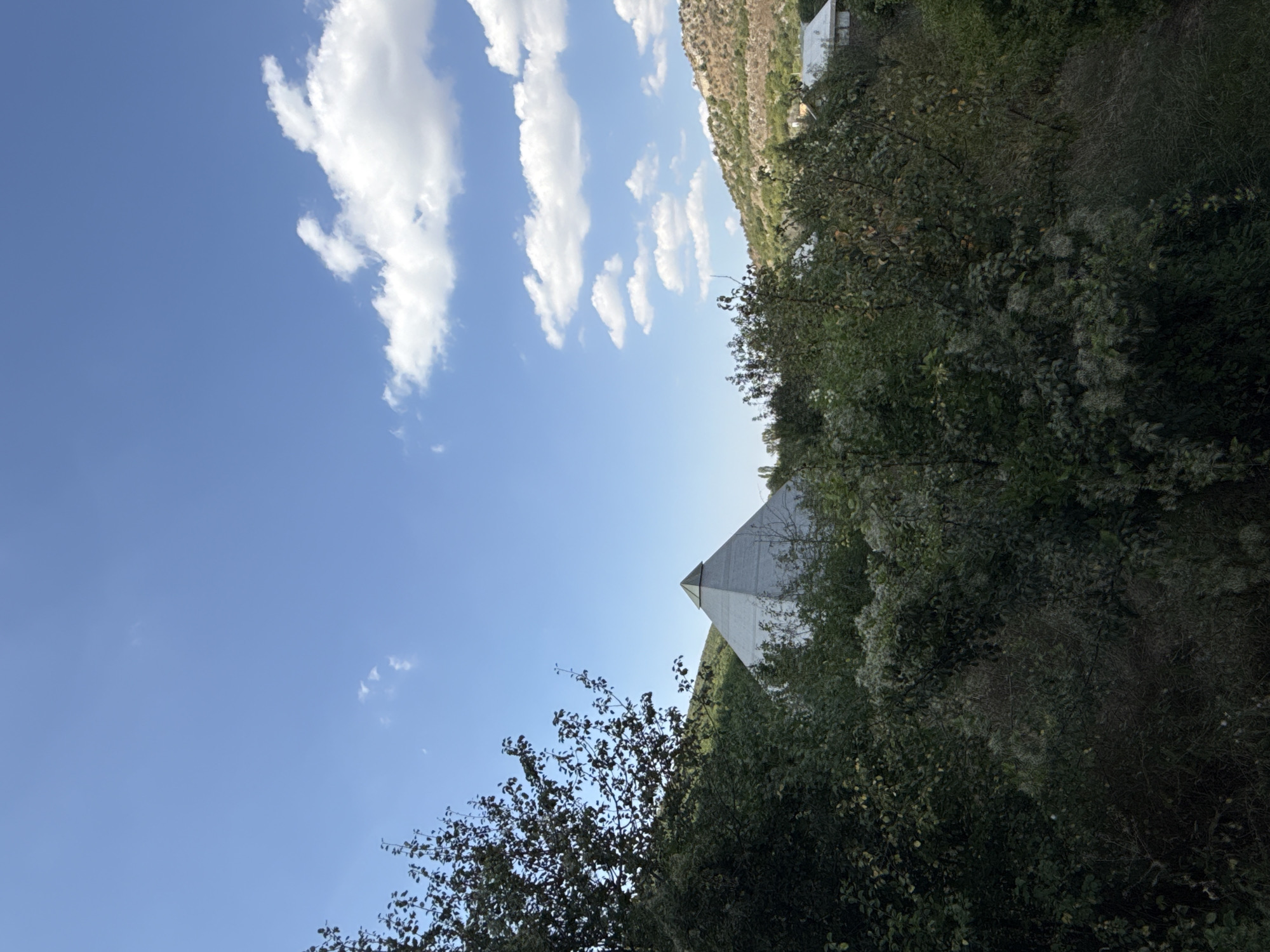 The image depicts a modern, pyramidal structure with a green apex, peeking out from behind lush green foliage. The building is situated in a hilly, somewhat overgrown landscape, with a clear blue sky dotted with white clouds overhead. The surrounding environment suggests a semi-rural or natural setting, with a few scattered buildings visible in the distance on the right side of the frame. The foreground is dominated by dense trees and bushes, partially obscuring the view of the pyramid. The lighting indicates it is daytime, likely mid-morning or afternoon, given the brightness and the presence of shadows. No people are visible in the image. The architecture of the pyramid suggests a contemporary design. The location is identified as the Скалистовское сельское поселение in Ukraine.