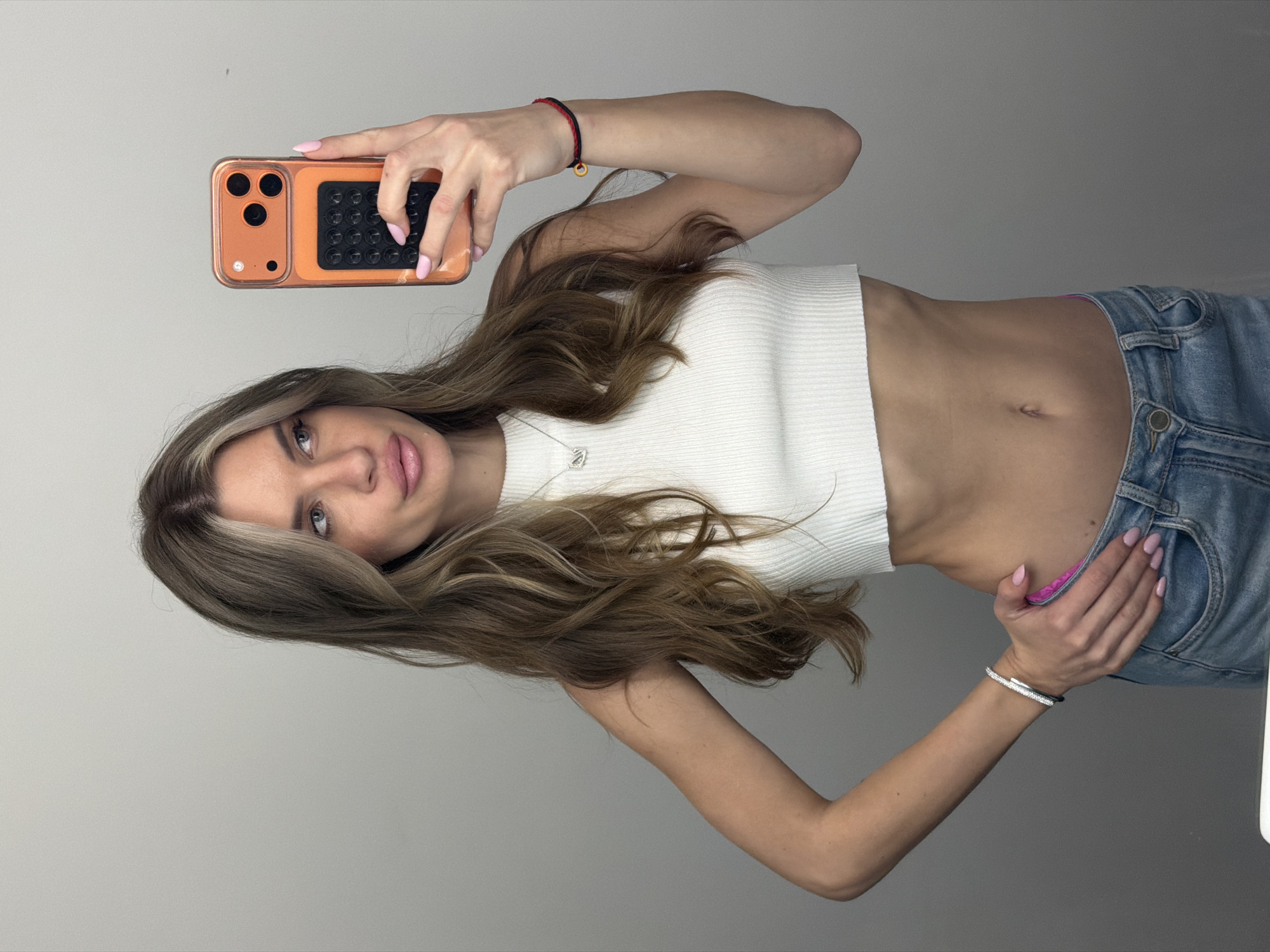 A woman with long, highlighted brown hair is taking a selfie with a smartphone. She's wearing a white cropped top and light-wash jeans. She is standing indoors against a plain, gray wall. The location is not identifiable as Berlin, Germany, based solely on the image.