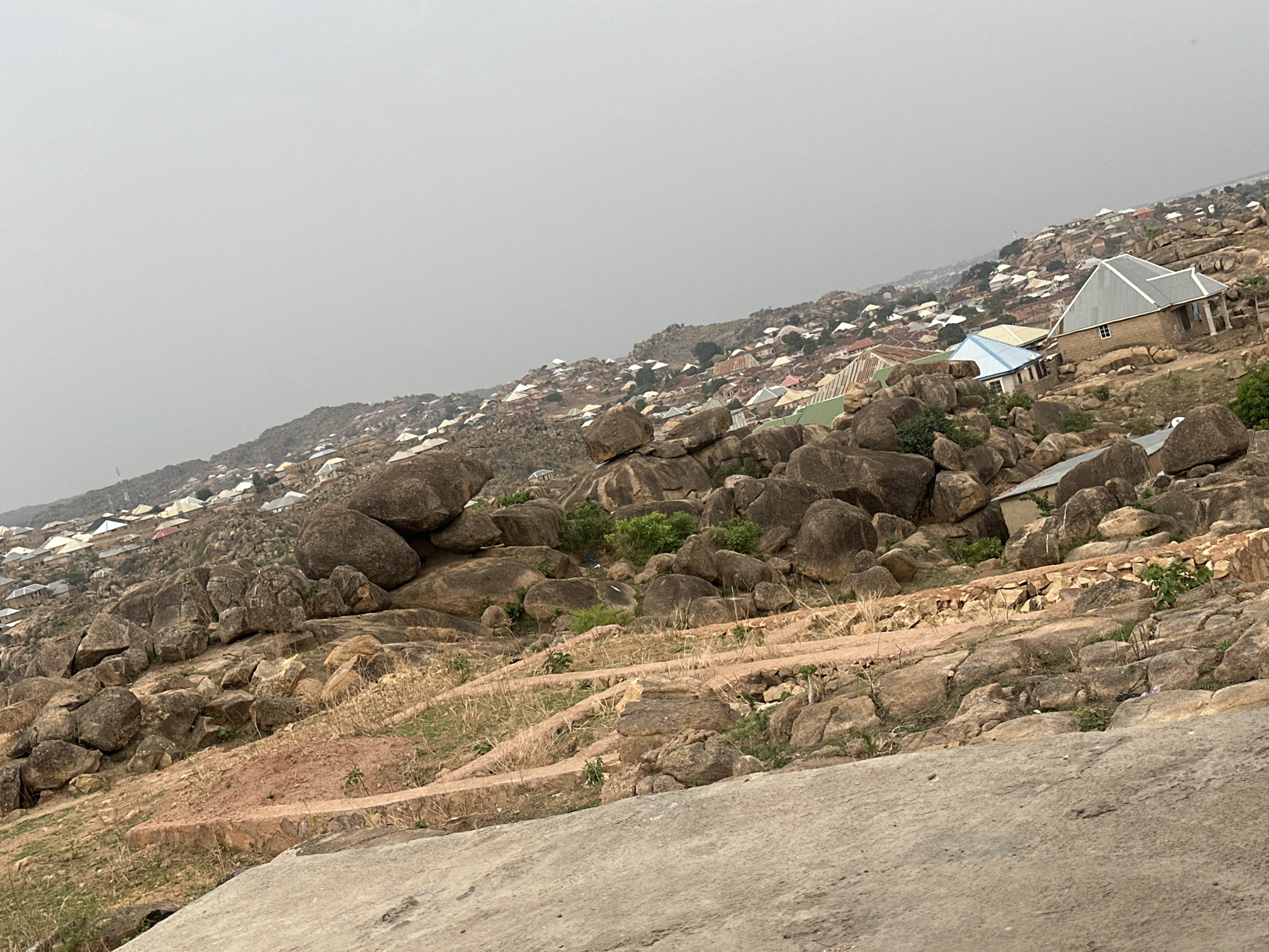 The image depicts a densely populated area in Jos, Nigeria, built on a rocky, uneven terrain. Houses of varying sizes and colors are scattered across the hillside, interspersed with large boulders and sparse vegetation. The sky is overcast, creating a muted, gray light. In the foreground, a concrete structure or surface is visible. The overall impression is of a challenging environment where settlement has adapted to the natural landscape.