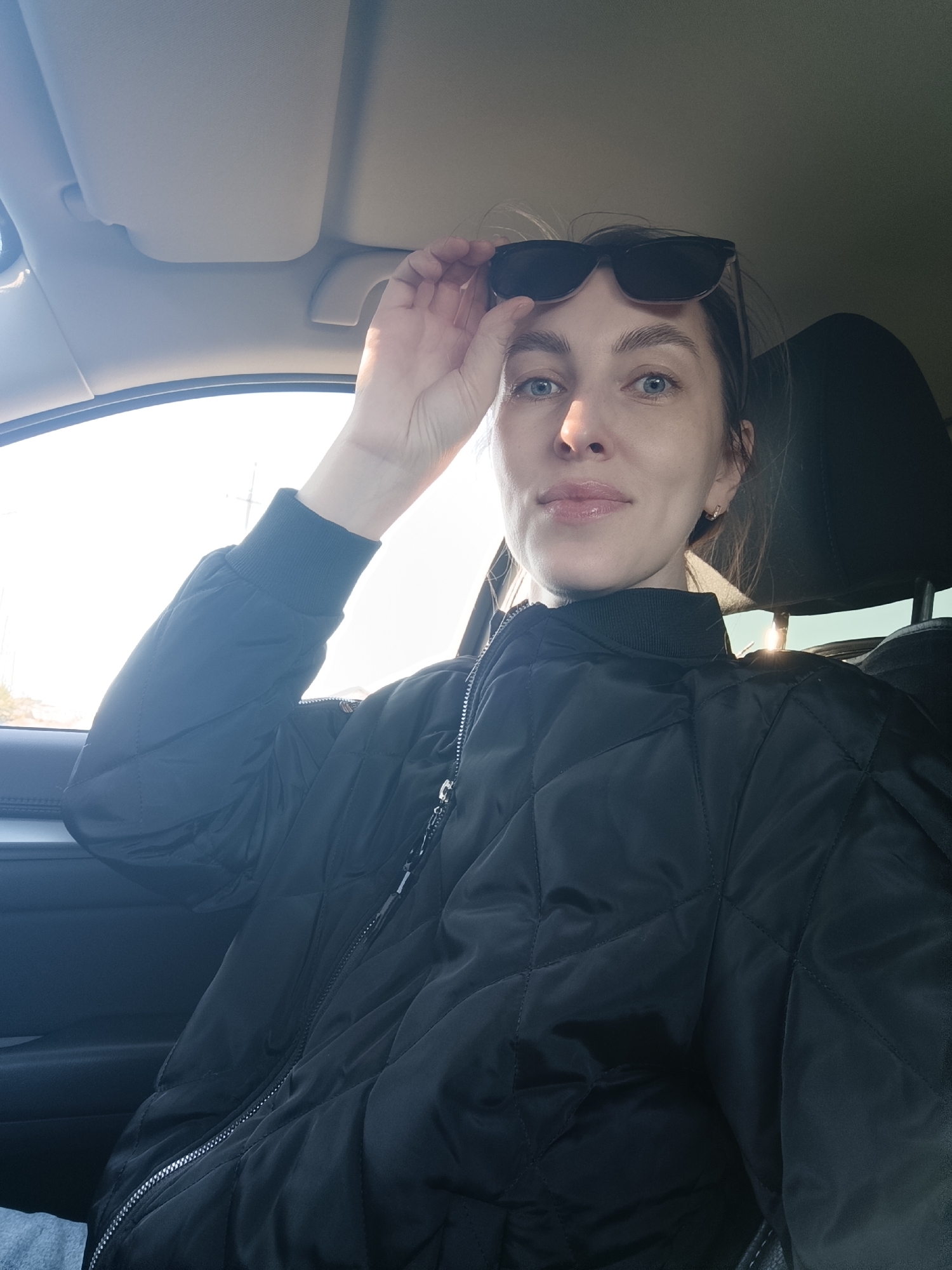A woman is sitting in the driver's seat of a car. She is holding her sunglasses above her eyes, looking at the camera. She is wearing a black quilted jacket. The sunlight is shining through the car window. The interior of the car is visible. There is no information to identify the specific location as Chaikovsky, Russia.