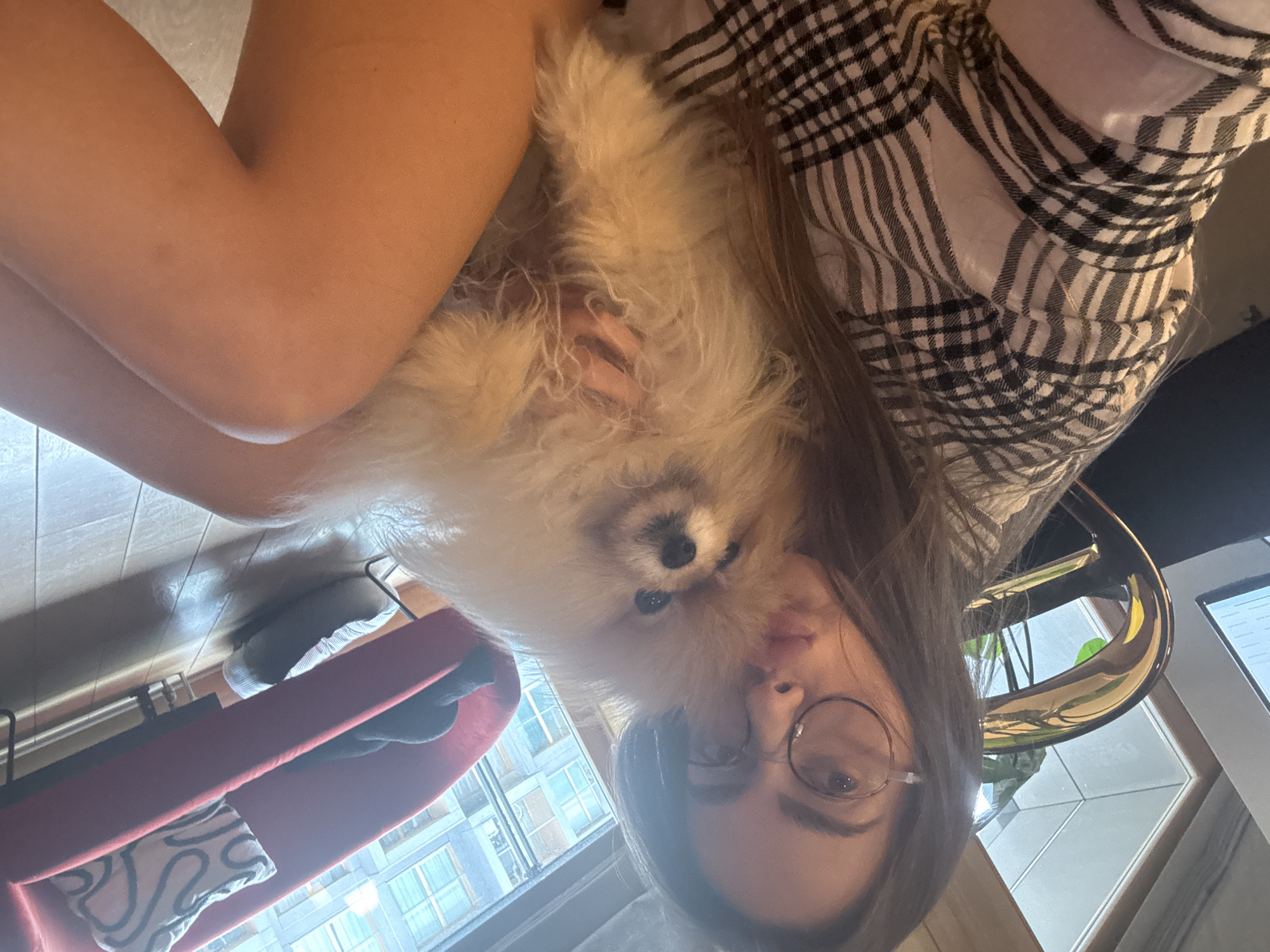 A rotated indoor selfie shows a woman with long brown hair and round glasses with a white fluffy dog. The woman is wearing a black and white checkered shirt. A red chair and a window with a view of buildings can be seen in the background. The location is potentially in Warsaw, Poland, based on the urban architectural style visible through the window.