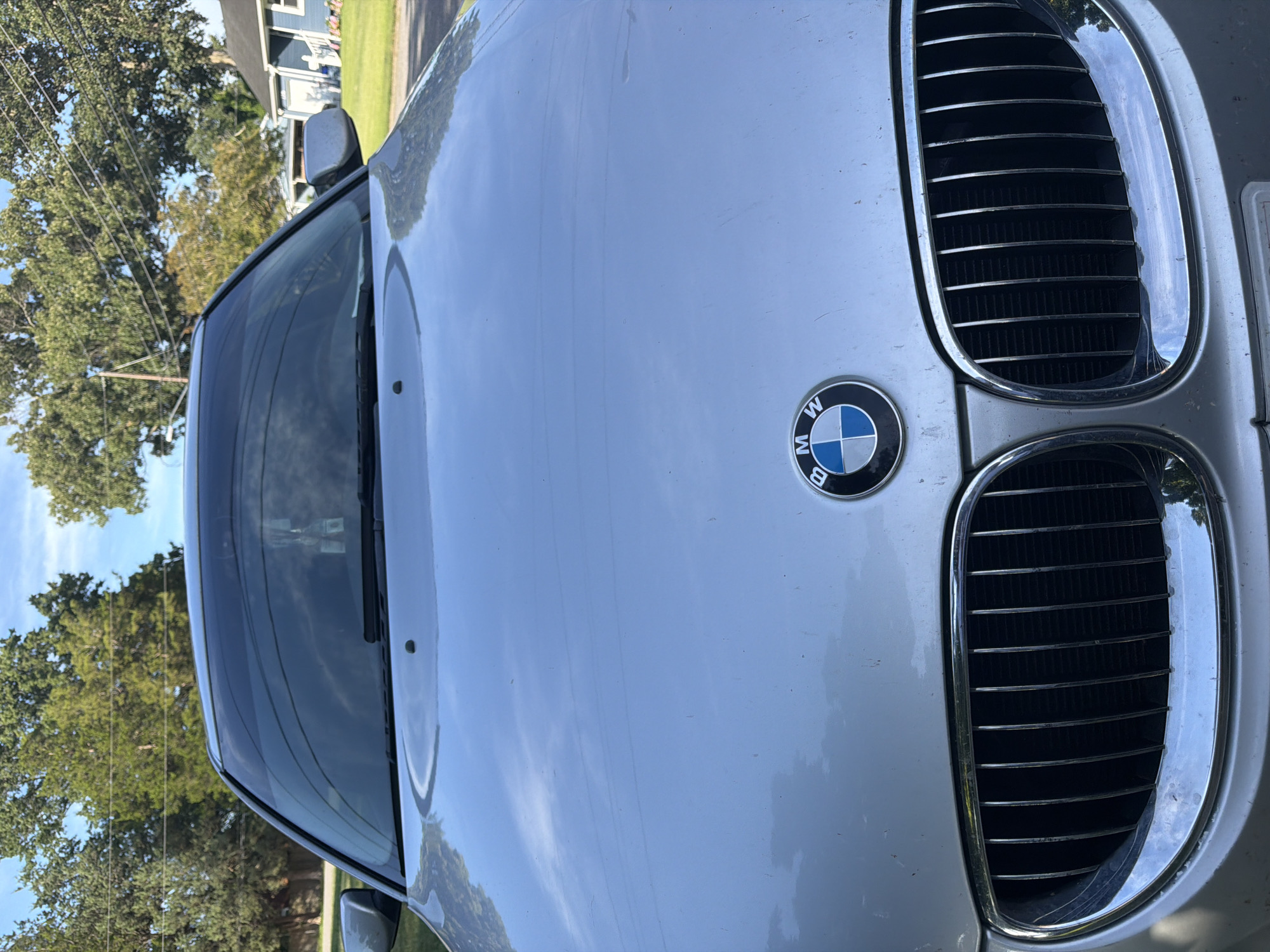 The image shows the front of a silver BMW car parked on a residential street in Virginia Beach, United States of America. The car is facing the camera, and the hood, grille, and windshield are visible. The BMW logo is prominently displayed on the hood. Trees and a house are visible in the background, suggesting a suburban environment. The sky is blue with some clouds.