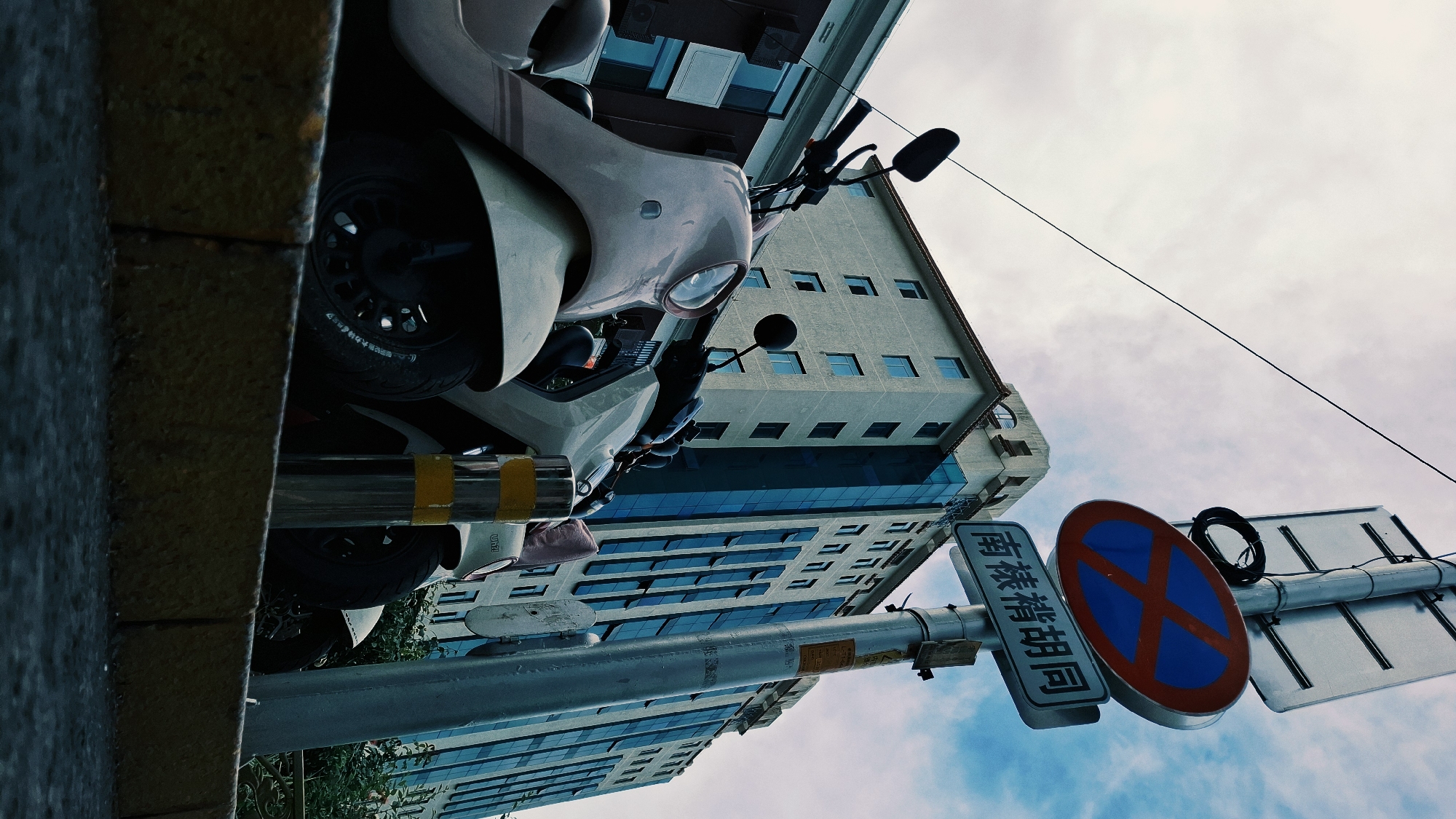 A low-angle, rotated shot captures a street scene in Dongcheng District, China. In the foreground, a parked cream-colored scooter is partially visible. A utility pole displaying a blue and red "no parking" sign with Chinese text is prominent. Behind the scooter and sign, a tall building with a glass facade and multiple windows fills the frame. The sky, partly cloudy, forms the backdrop. The image is taken from a position close to the ground.