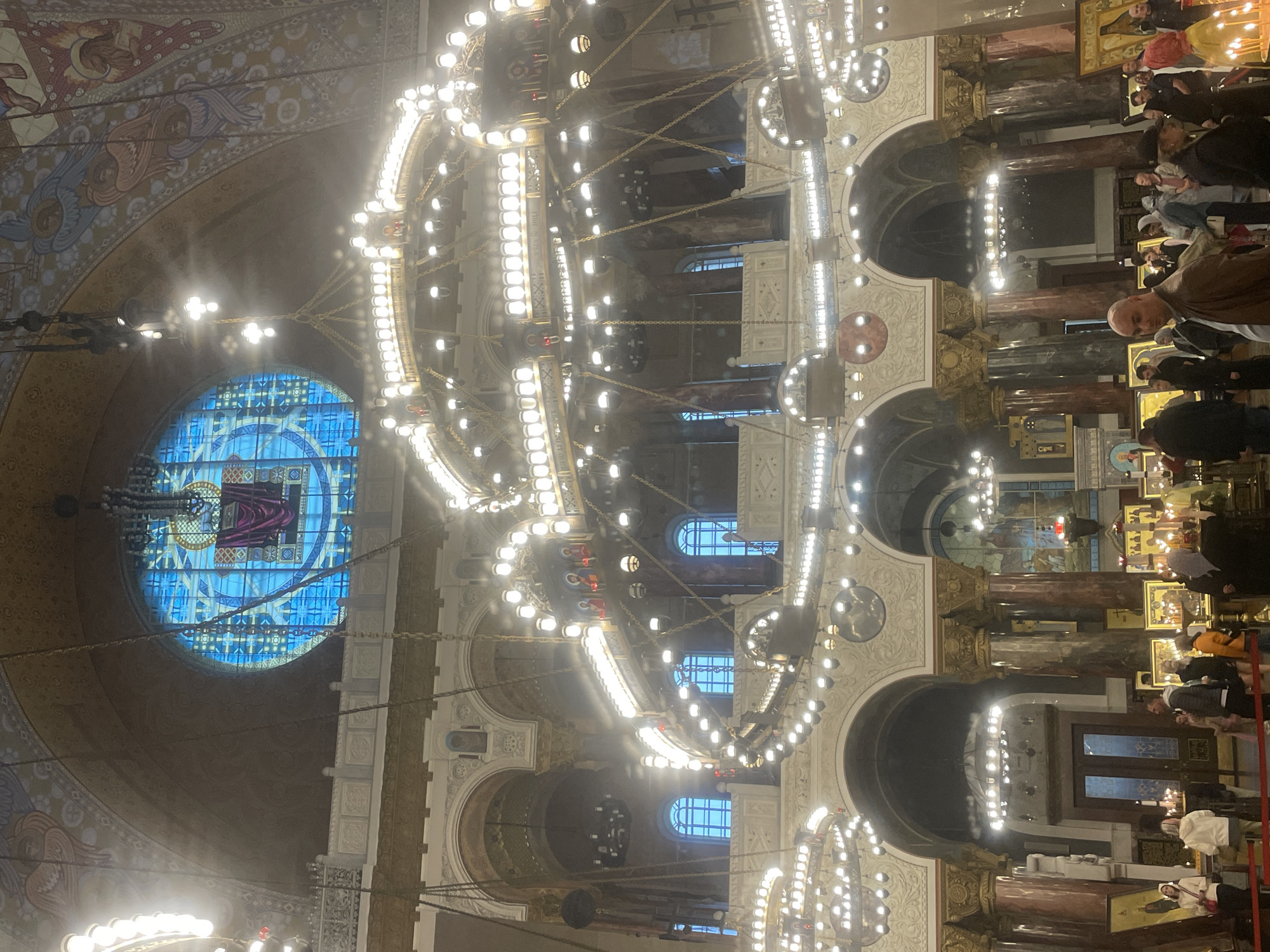 The image shows the interior of a large, ornate building, likely a cathedral or church, in Kronstadt, Russia. The architecture features soaring arches, decorative moldings, and richly colored murals, including depictions of angels on the upper walls. Large, multi-tiered chandeliers with numerous lights hang from the ceiling, illuminating the vast space. A prominent circular stained-glass window with a blue hue and religious imagery is visible in the upper part of the frame. Below, several smaller windows with the same blue tint are set into the upper level. The lower level of the building has a grand entrance with double doors and is supported by large, marble-clad columns. Along the walls, various religious icons and paintings are displayed. A crowd of people, dressed in casual attire, are gathered inside, some looking at the artwork, others engaged in conversation. The overall atmosphere appears to be one of reverence and admiration for the grandeur of the religious edifice.