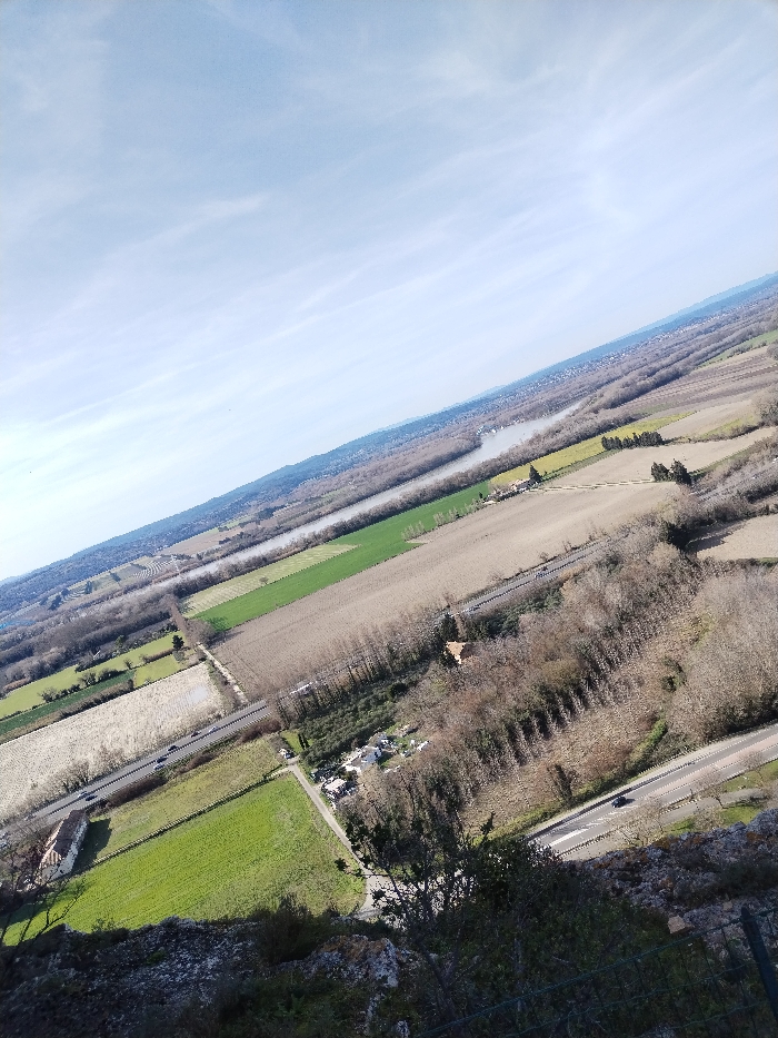 The image captures a vast, tilted aerial or high-vantage-point landscape view on a bright, clear day, likely late autumn or winter, given the bare deciduous trees. The perspective is looking down and across a wide valley or plain, with a slight diagonal tilt to the entire scene, making the horizon line appear sloped.
The top portion of the image is dominated by a pale blue sky, streaked with delicate, wispy white clouds that extend across the frame. Below the sky, the distant horizon reveals a low range of hills or mountains, appearing hazy and soft.
A prominent river or large body of water snakes its way through the mid-ground, reflecting the sky and appearing a light, silvery gray. It flows generally from the upper right towards the mid-left of the image. Along its banks and spreading throughout the valley are extensive areas of land segmented into various fields. Some fields are vibrant green, suggesting active agriculture or pastureland, while others are a lighter, muted beige or brown, indicative of fallow land, harvested crops, or fields awaiting planting.
Interspersed among the fields and along the river are numerous clusters and lines of trees. These trees are mostly bare of leaves, showing the skeletal structure of their branches, which further suggests a colder season. Some denser wooded areas form dark patches or strips across the landscape.
Human infrastructure is visible throughout the scene. A main paved road, with clear white lane markings, cuts diagonally from the bottom right quadrant of the image towards the mid-left, paralleling some of the fields and tree lines. Several vehicles, appearing as small dark shapes, can be seen traveling on this road. Another, narrower road or track, possibly unpaved, is visible further to the left, connecting different parts of the farmland. A railway line, distinguished by its straight, dark path, also runs through the landscape, generally parallel to the river and some of the fields.
Scattered buildings dot the landscape. A notable structure with a reddish-brown roof and light-colored walls stands near the bottom-left, adjacent to a green field and a road. Other smaller buildings or faint clusters of habitation can be observed in the distance, particularly along the riverbanks.
The immediate foreground, at the very bottom of the image, shows the rocky and somewhat rugged terrain from which the photograph was taken. This area is composed of rough, light-colored rocks and sparse, dark green foliage, including bushes and small trees. A faint, dark, wire-like fence structure is barely visible at the extreme bottom right corner, suggesting a barrier at the elevated viewpoint.
The overall impression is one of a vast, tranquil, and agricultural landscape, viewed from a considerable height under bright daylight. There is no visible text in the image.