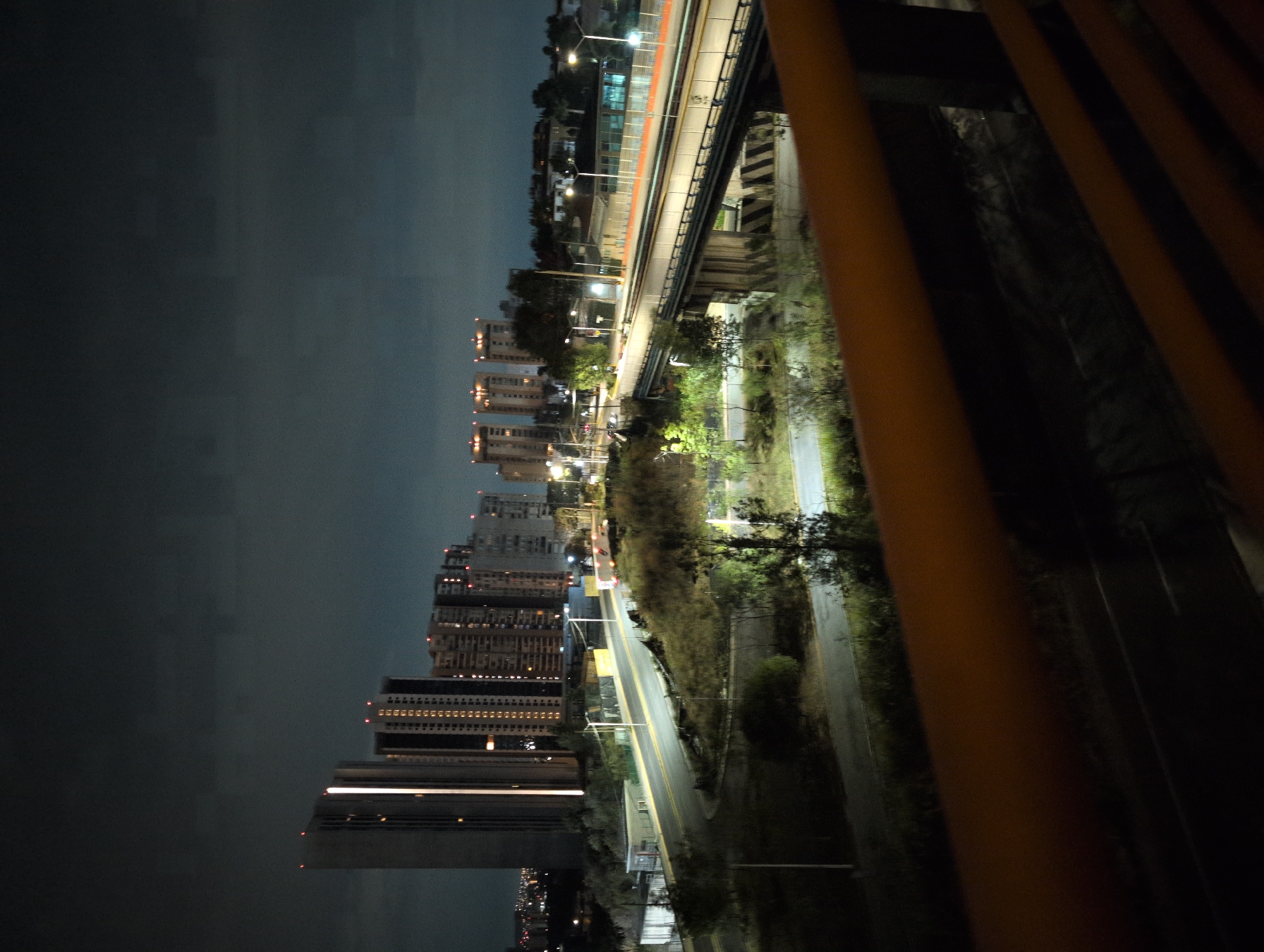 The image shows a nighttime urban scene in Jesús del Monte, Mexico, possibly taken from an elevated position such as a bridge or building. A row of tall buildings lines the horizon on the left, illuminated with numerous lights. A road with streetlights runs parallel to the buildings. An elevated railway or highway structure, also lit, extends across the top portion of the image. The right side of the image is obscured by a series of vertical orange bars, possibly a railing. Vegetation is visible between the road and the elevated structure. The sky is dark, suggesting late evening or night. No people are visible.