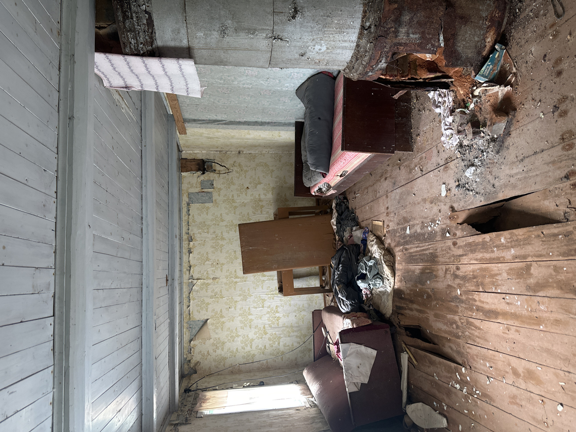 This image depicts the interior of a derelict room in an abandoned building, likely in the Відзаўскі сельскі Савет, Belarus. The room is in a state of disrepair, with peeling wallpaper, damaged wooden floors, and exposed ceiling beams. A sleeping bag is laid out on a mattress atop a wooden structure, suggesting a makeshift living space. Scattered debris, including torn fabric, paper, and what appears to be building materials, litters the floor and a hole in the ceiling. A small wooden table and a dark red armchair are also present, further indicating past habitation. The overall impression is one of neglect and decay. The lighting suggests it is daytime, with natural light entering through a window on the left side of the frame. There is no visible text in the image.