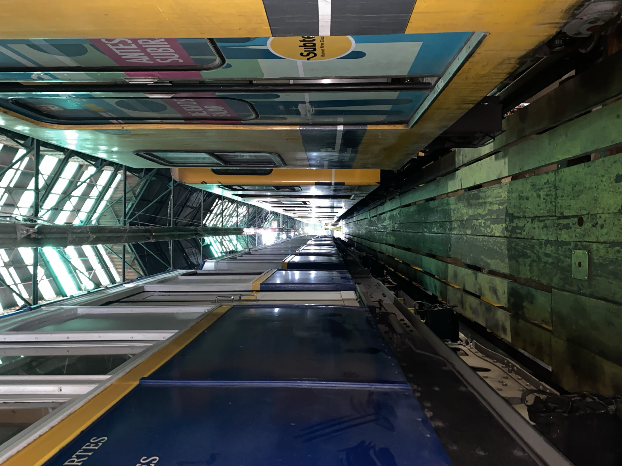 The image depicts a train car of the Subte (subway) system in Buenos Aires, Argentina, inside a workshop or maintenance depot. The train car is positioned between a green wooden platform or structure on the right and the steel framework of the workshop on the left. The top of the train car is blue and yellow with the words "Antes Subir". The other car has yellow paint and a "Subte Buenos Aires Ciudad" logo. The perspective is looking down the length of the train, which fades into the distance. The angle suggests that the photo was taken from above the car with the camera rotated, creating a slightly disorienting perspective. The environment is industrial, with green-painted wooden structures on the right and visible daylight from the open-air workshop or maintenance depot.