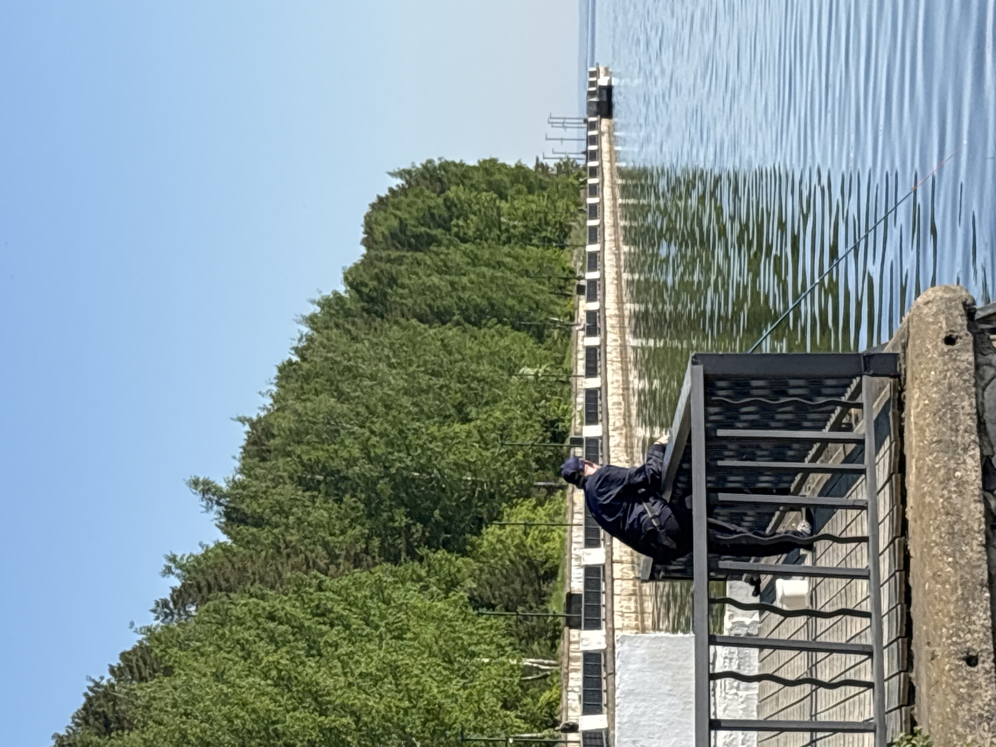 A person wearing a dark blue jacket and cap is fishing from a metal staircase on the edge of a pier or dock. The body of water is calm with wave patterns. The pier appears to be located in the city of Chaikovsky, Russia. The location is adjacent to a densely forested area. The sky is clear and blue.