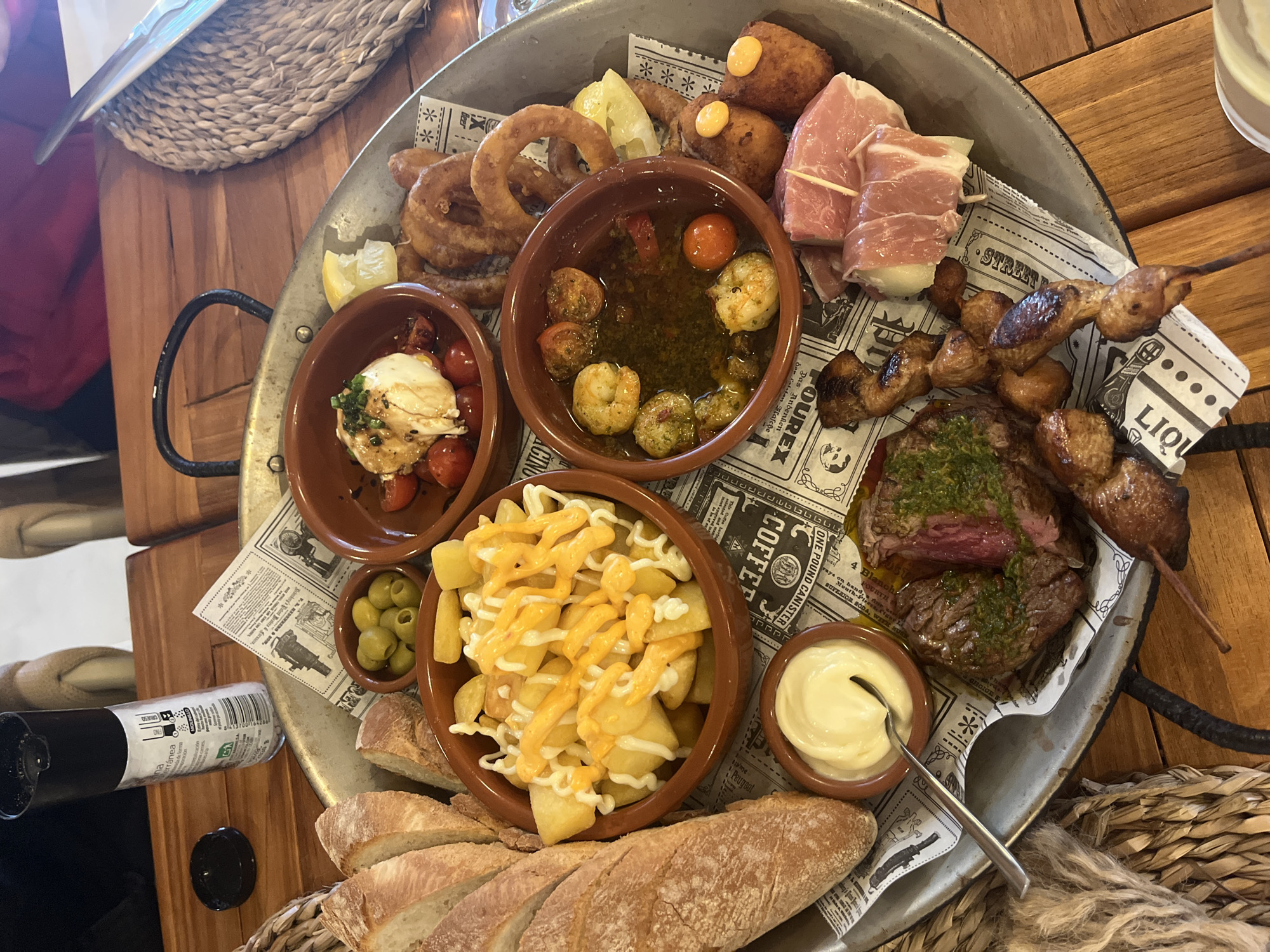 The image captures a large, round, metal platter brimming with various tapas. The platter is lined with newspaper-themed paper.
Food items include: onion rings, deep-fried croquettes with a yellow sauce, prosciutto-wrapped bites, a small bowl with shrimp in sauce, grilled chicken skewers, steak topped with green sauce, a bowl of fried potatoes with yellow and white sauce, olives, bread, and a small bowl of mozzarella and tomatoes. A small bowl of mayonnaise and a serving spoon are also visible.
A black bottle is lying on the table to the side of the tray. The table itself appears to be made of light-colored wood. A woven basket-like object is also present, likely serving as a place mat or decorative element.
The location appears to be a restaurant, presumably in Xàbia / Jávea, Spain, based on the traditional Spanish tapas served.