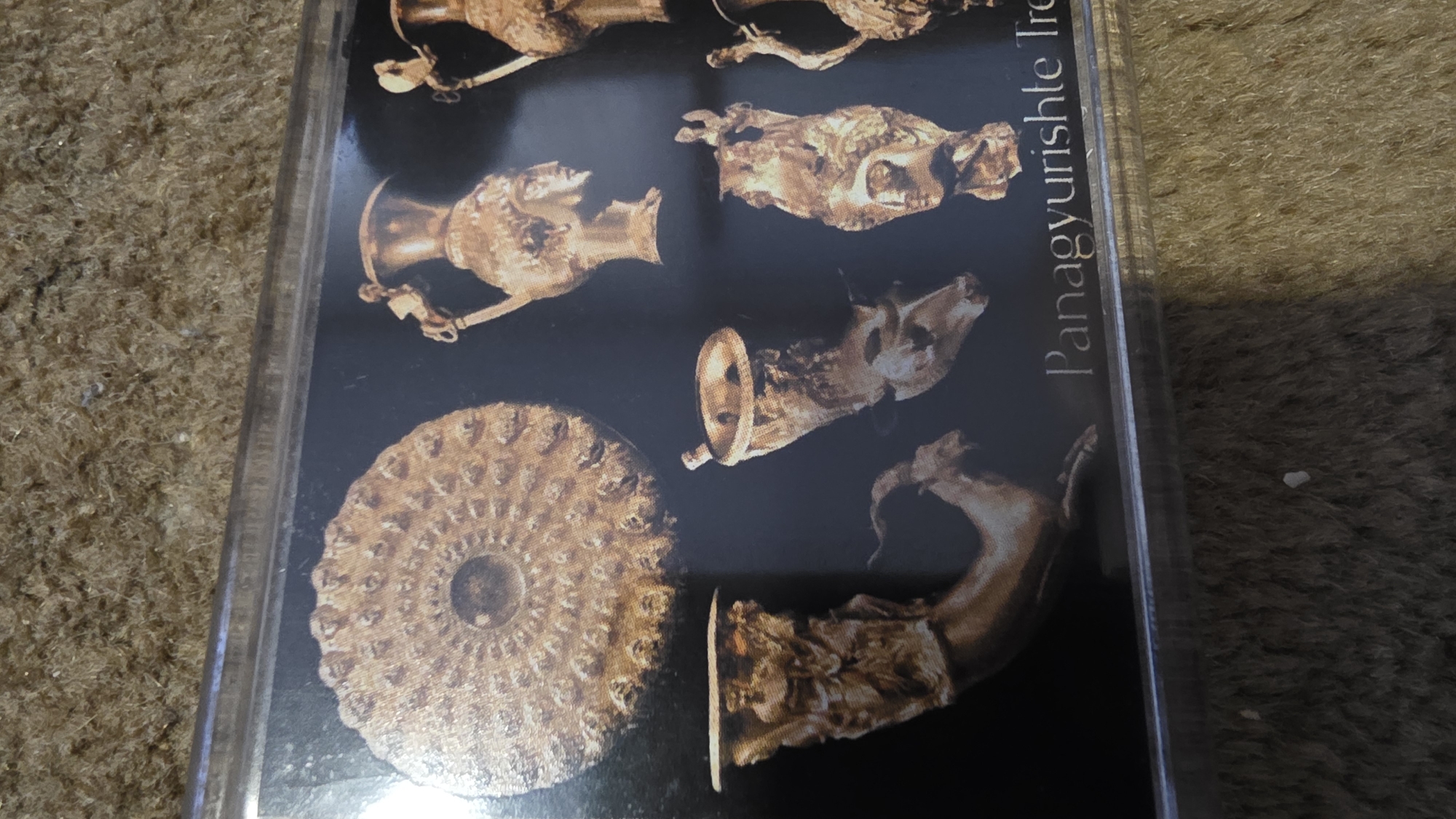 The image shows a framed photograph or print of artifacts from the Panagyurishte Treasure. The print includes images of various golden vessels, including rhytons (drinking horns shaped like animal heads), and a flat, round object with intricate concentric patterns. The text "Panagyurishte Tre" is visible on the right side of the print. The background of the photograph is black. The framed print appears to be resting on a surface covered with brown, possibly carpeted, material. While the location is not explicitly stated, the Panagyurishte Treasure is associated with the region around Panagyurishte, Bulgaria, and exhibits of the treasure are sometimes located in Blagoevgrad, Bulgaria.
