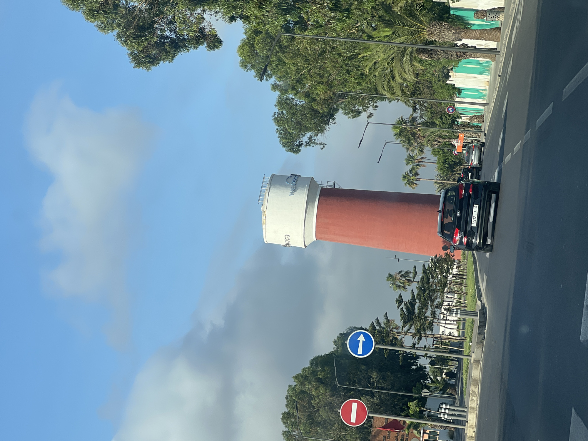In Casablanca, Morocco, a wide street is visible with cars driving on it, including a black Hyundai i30. On the left side of the street, there are two traffic signs: a red circular sign with a horizontal white bar and a blue circular sign with a white right arrow. Beyond the signs, there is a tall red and white water tower with the logo "WE" on it, surrounded by trees and palm trees. On the right side of the street, there is a green and white striped wall with palm trees and streetlights lining the sidewalk. The sky is blue with some clouds.
