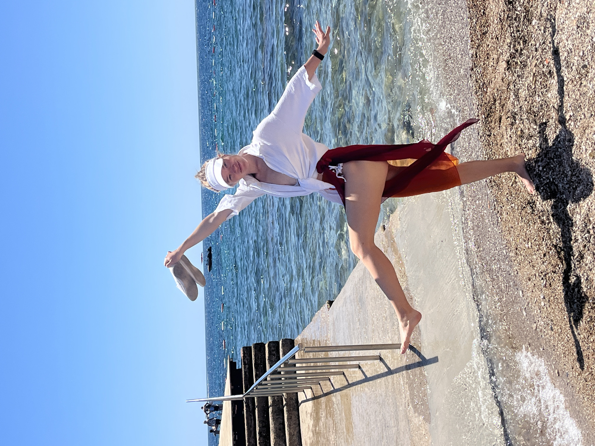 A photograph depicts a woman in a dynamic pose on a pier in Budva, Montenegro. She is barefoot, wearing a white open shirt, and a reddish-orange sarong-like garment. A white visor is on her head. She is holding a pair of shoes in her right hand. Her arms are extended. The pier has a metal railing and steps. The sea is visible in the background, with clear blue water meeting the pier. To the right of the pier is a beach with beige sand or pebbles. The sky is a clear blue. The photo has been rotated.