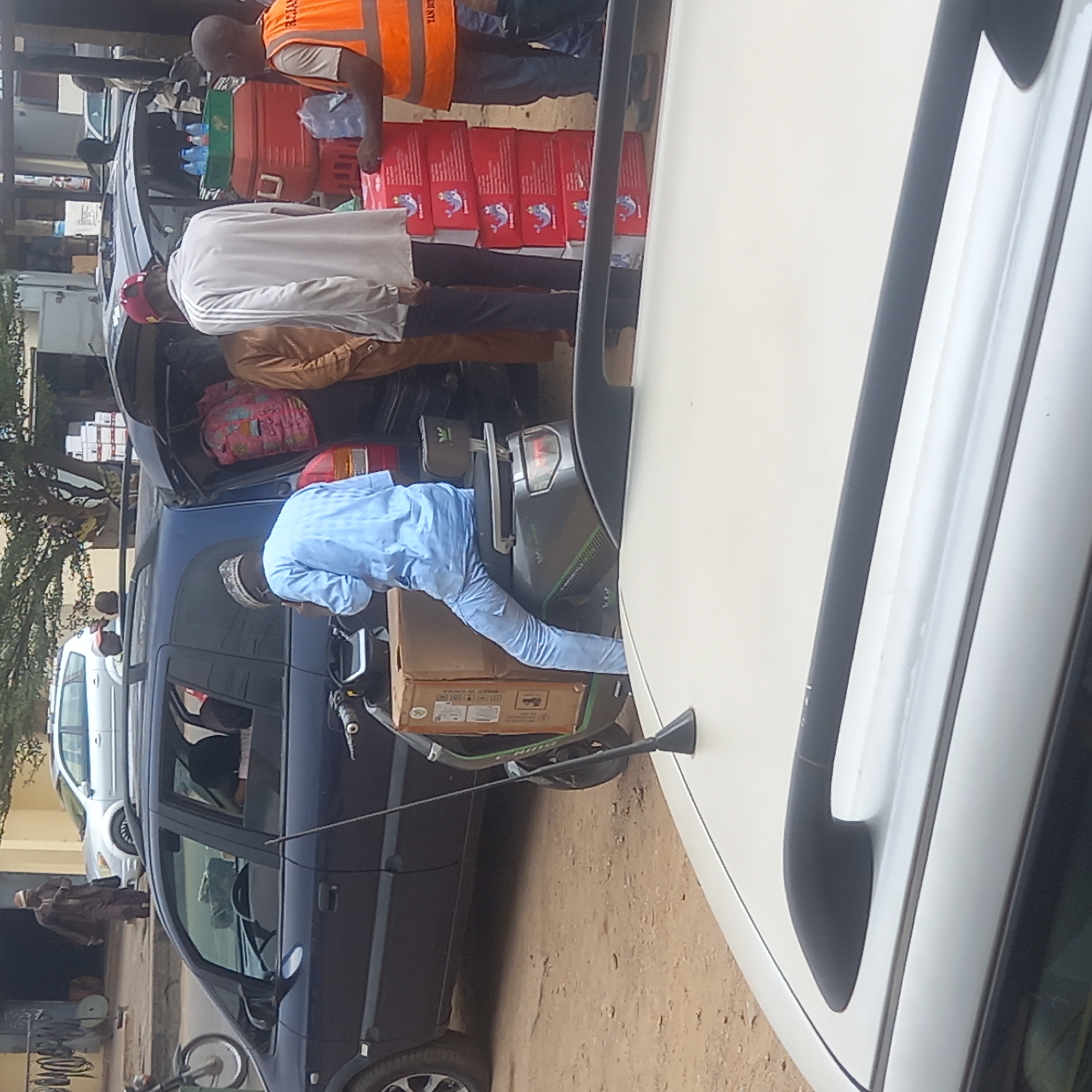 The image depicts a street scene in Dadi Riba, Nigeria. People are unloading items from a blue car and a black car, potentially supplies or goods. Red boxes with a blue and white logo and other items are stacked on the black car. A man in an orange vest is standing beside the stacked items. A man in a blue shirt is placing a cardboard box on a scooter. Other people are present in the background. The environment appears to be a street with buildings and other vehicles.