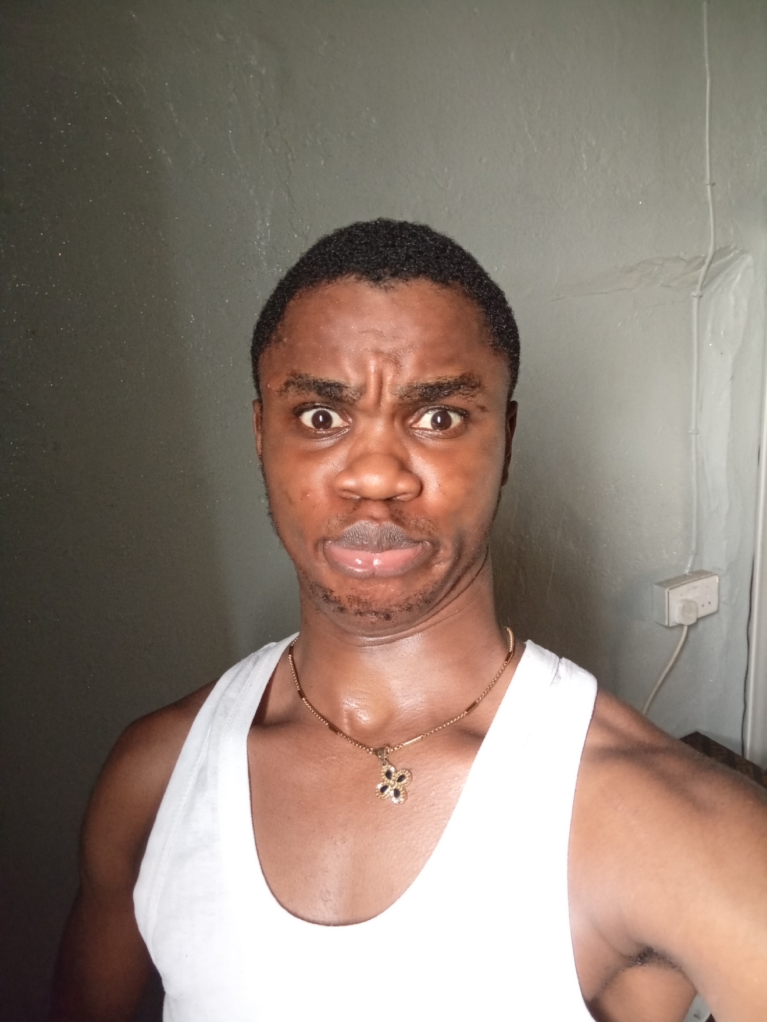 A medium shot captures a man with brown skin, a dark, short haircut, and a gold necklace with a flower pendant. He is wearing a white tank top. His facial expression is characterized by furrowed brows, wide eyes, and a puckered mouth. The background is a plain, grayish wall with what appears to be an electrical outlet on the right side. The location is Unknown, Unknown.
