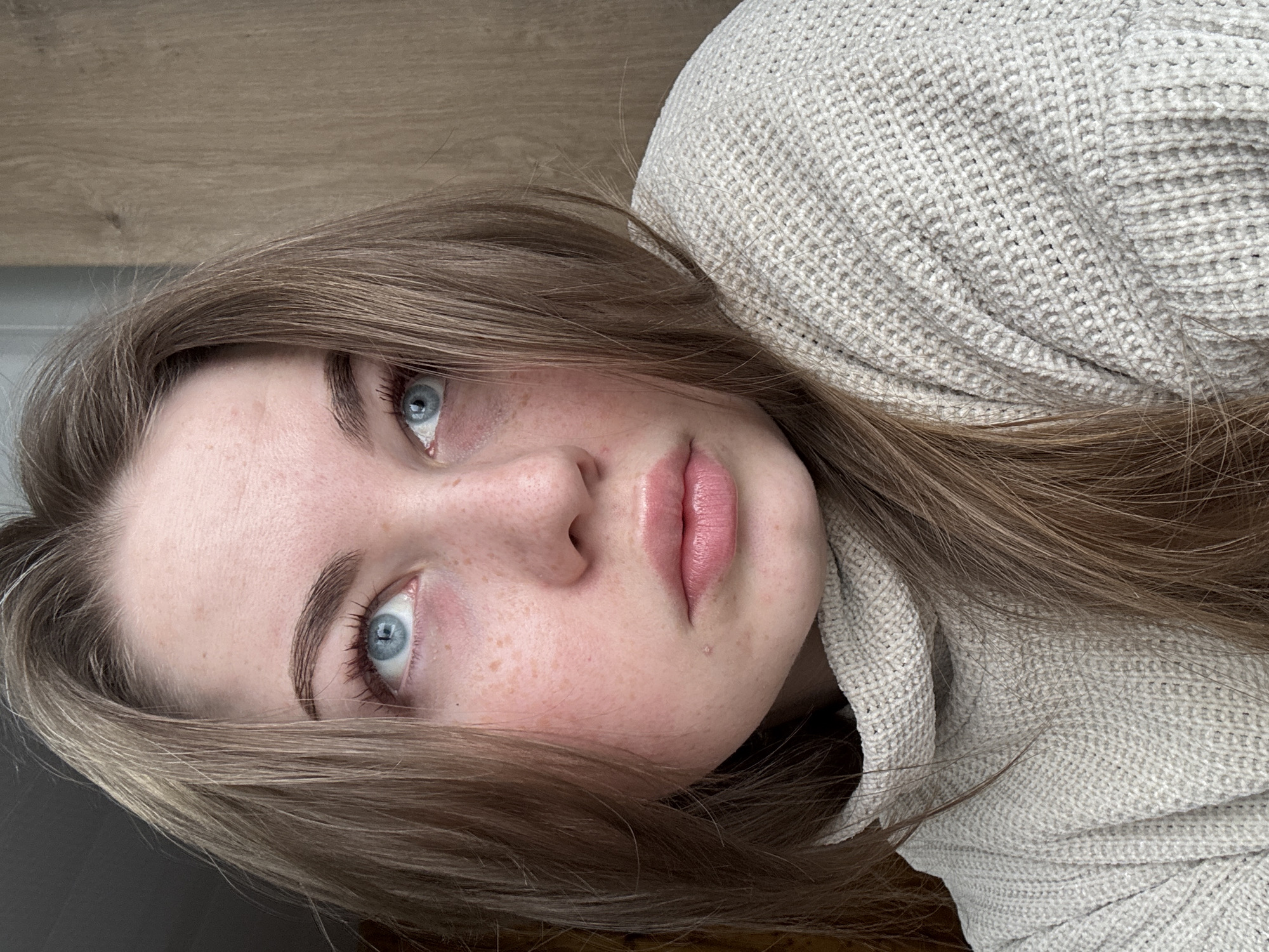A close-up, rotated portrait features a young woman with light brown hair and blue eyes. She has a fair complexion with freckles visible on her cheeks and nose. Her lips are full and pink. She is wearing a light-colored, textured sweater. The background consists of a light brown wooden surface.
I am unable to verify the city of Уральское, Russia.