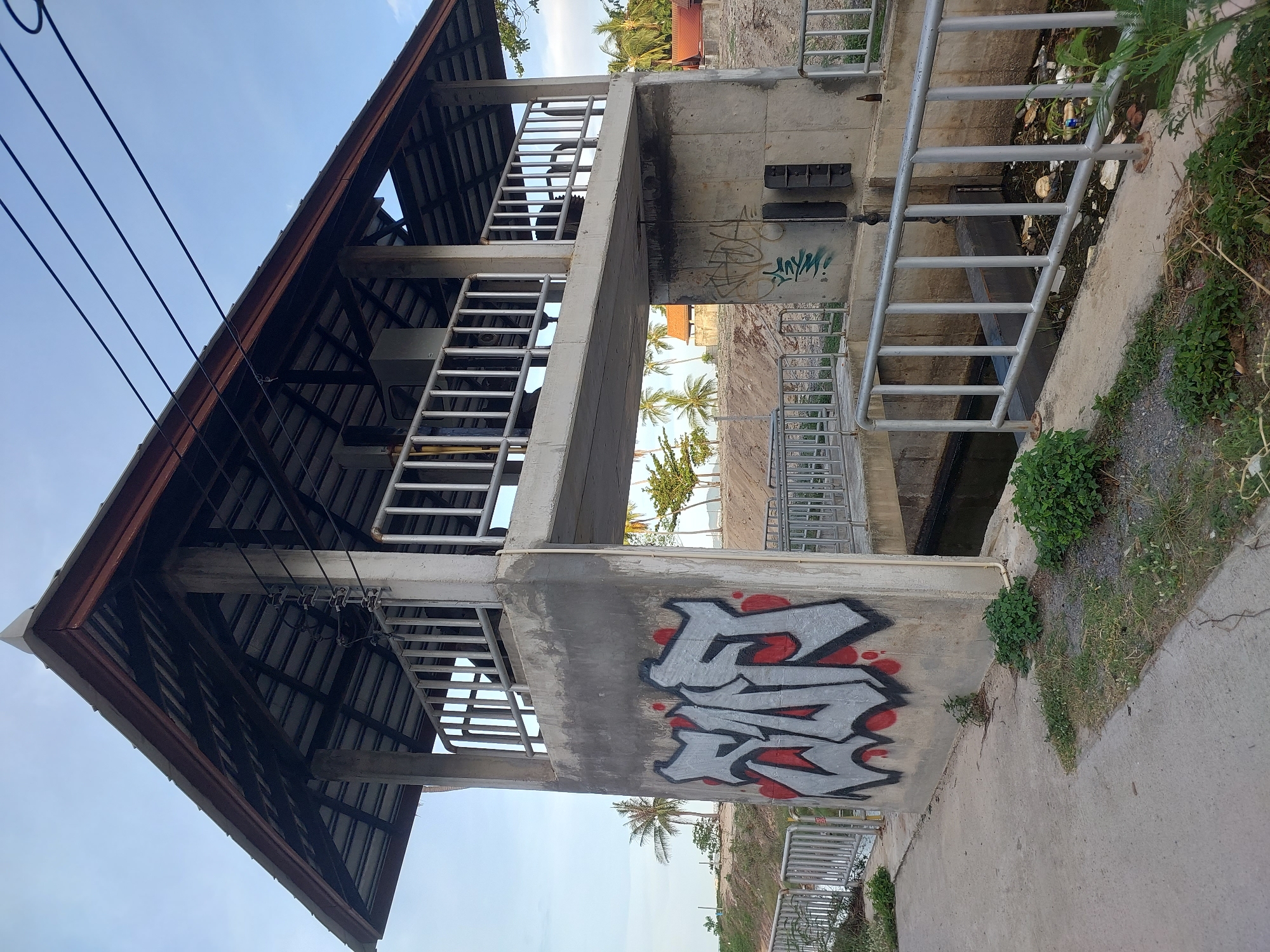 This is an eye-level, slightly tilted, outdoor shot of a multi-level concrete structure with a metal roof in Baan Tai, Thailand. The structure appears to be a kind of covered walkway or viewing platform, with metal railings and stairs. It has a utilitarian design, with exposed concrete and metal supports.
Graffiti is visible on the lower concrete wall, featuring bold, stylized lettering in grey with red outlines. The surrounding environment suggests a tropical location, with palm trees visible in the background and some vegetation growing around the base of the structure. The ground is a mix of concrete and gravel. A narrow channel of water, appearing somewhat polluted with debris, runs alongside the structure, bordered by metal railings. Overhead, power lines are visible against a clear blue sky, indicating it is daytime with fair weather. The scene feels somewhat neglected, with the graffiti and the debris in the water suggesting a lack of maintenance.