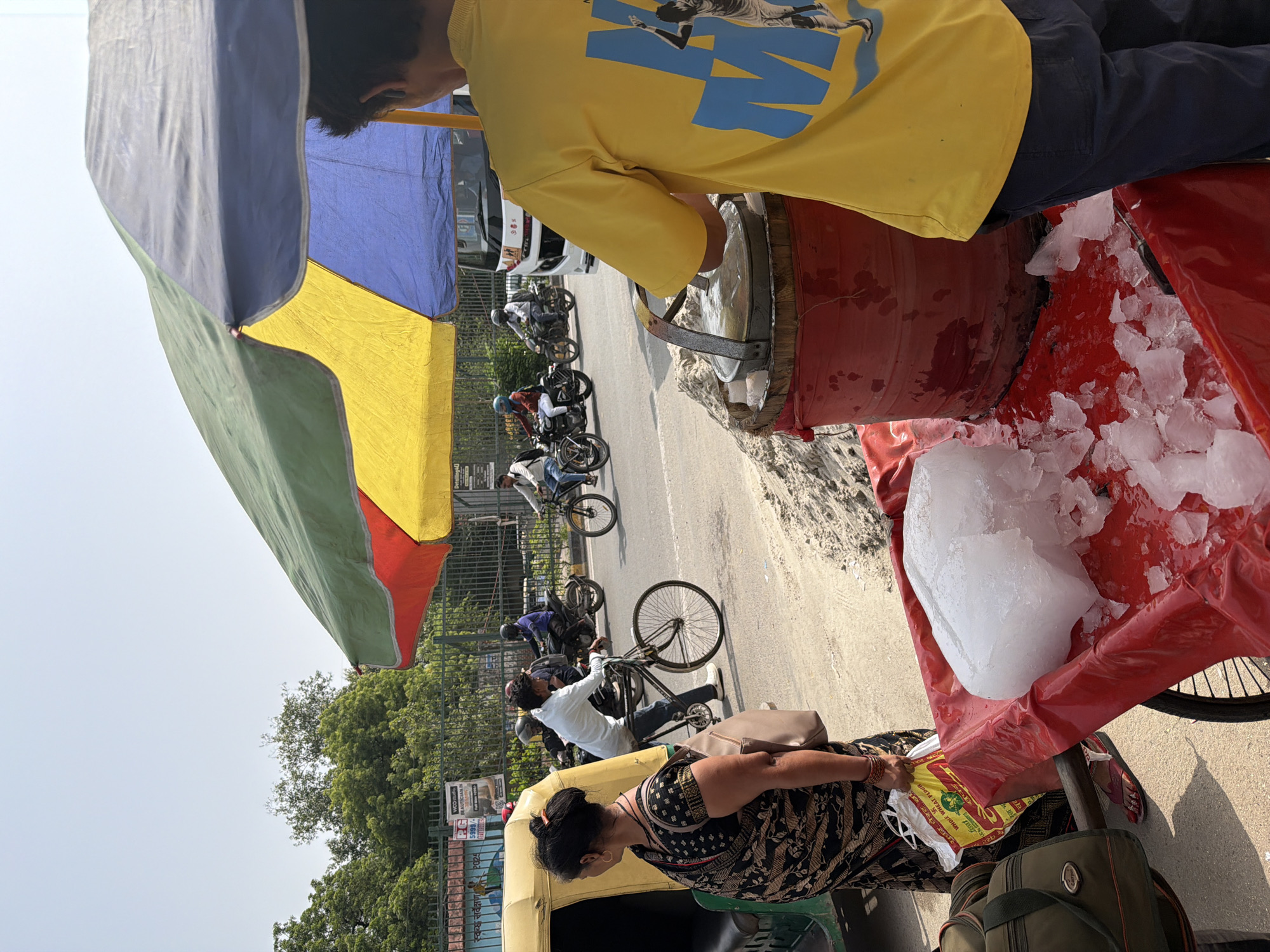 In Delhi, India, on a sunny day, a street vendor stands behind a cart laden with ice. The cart is covered with a bright red cloth, and a large block of ice sits prominently on top, with smaller pieces scattered around. The vendor, wearing a yellow t-shirt with blue graphics and dark pants, appears to be preparing to serve a customer or is in the process of selling ice. In the background, a busy street scene unfolds. An auto-rickshaw is parked on the left side of the image, and several people are riding motorcycles and bicycles. A bus is visible further down the road. The environment suggests a typical bustling street in Delhi, with buildings and trees lining the background. A large, multi-colored umbrella, red, yellow, and green, shades the vendor and the cart, indicating a public space where vendors often set up shop. The presence of the auto-rickshaw, the attire of the people, and the general atmosphere strongly indicate a location within the city of Delhi.
