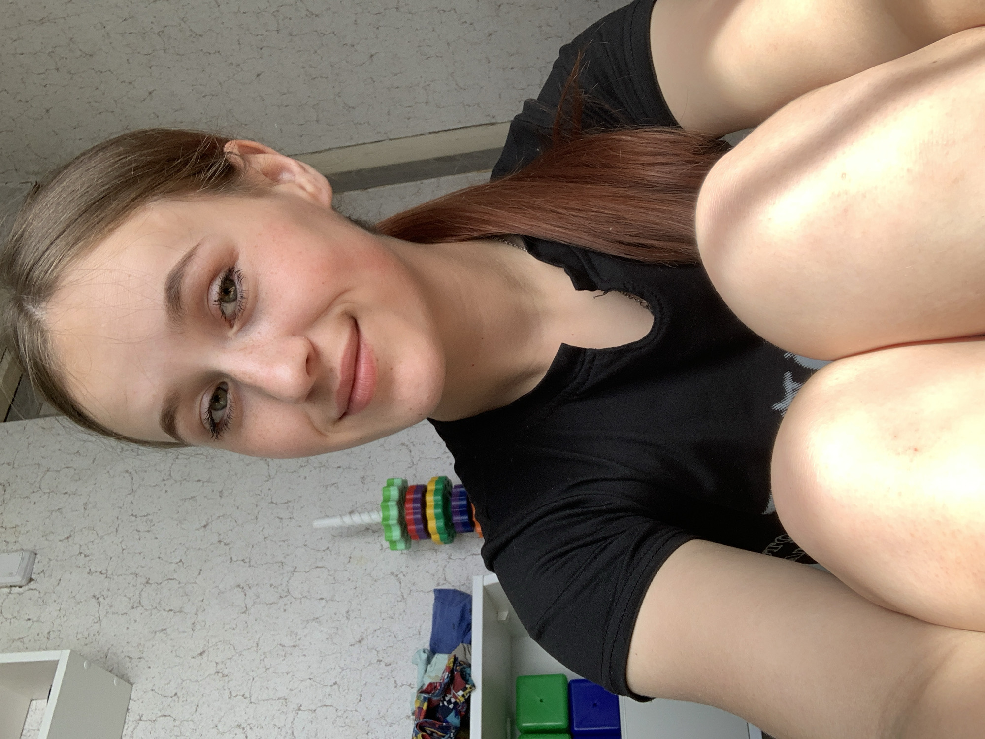 A woman with brown hair is looking at the camera with her legs up. She is wearing a black t-shirt. Behind her are toys. The location cannot be determined from the image.