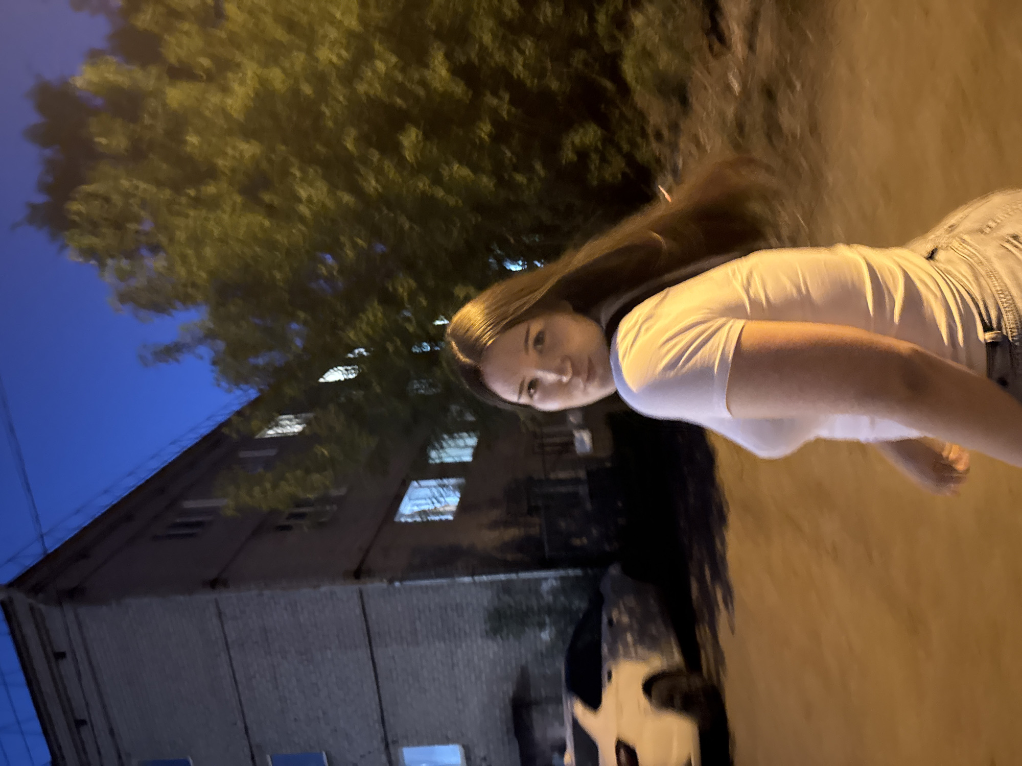 A young woman with long brown hair stands outdoors, facing the camera, in what appears to be the city of Perm, Russia. She wears a white t-shirt and denim shorts. Behind her is a multi-story brick building with illuminated windows, suggesting it is nighttime. A large tree with dense foliage is visible to the left and above the building, and a car is parked nearby. The lighting is mixed, with both natural and artificial sources illuminating the scene.