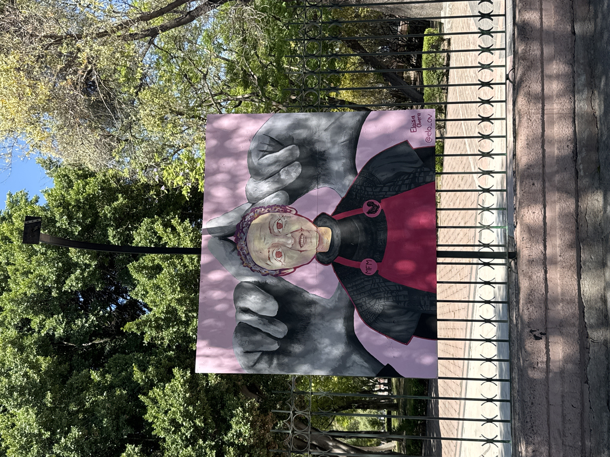 A photograph shows a street scene in Querétaro, Mexico. The image focuses on a large, square painting attached to a wrought-iron fence. The painting depicts a stylized portrait of a person with red eyes, set against a pink background. Overlapping the portrait are two large, gray-toned hands, positioned as if cradling or protecting the face. The portrait subject wears a dark-colored garment with a red bib-like section and a decorative button labeled "MFM." A small signature or attribution, "Elisa Olvera @elo_ov," is visible in the upper right corner of the painting.
The fence runs parallel to a sidewalk or paved area. Beyond the fence and painting is lush green foliage consisting of trees and bushes. The leaves suggest a warm climate and likely an urban park setting. The lighting indicates it is daytime, with sunny conditions casting shadows on the sidewalk.