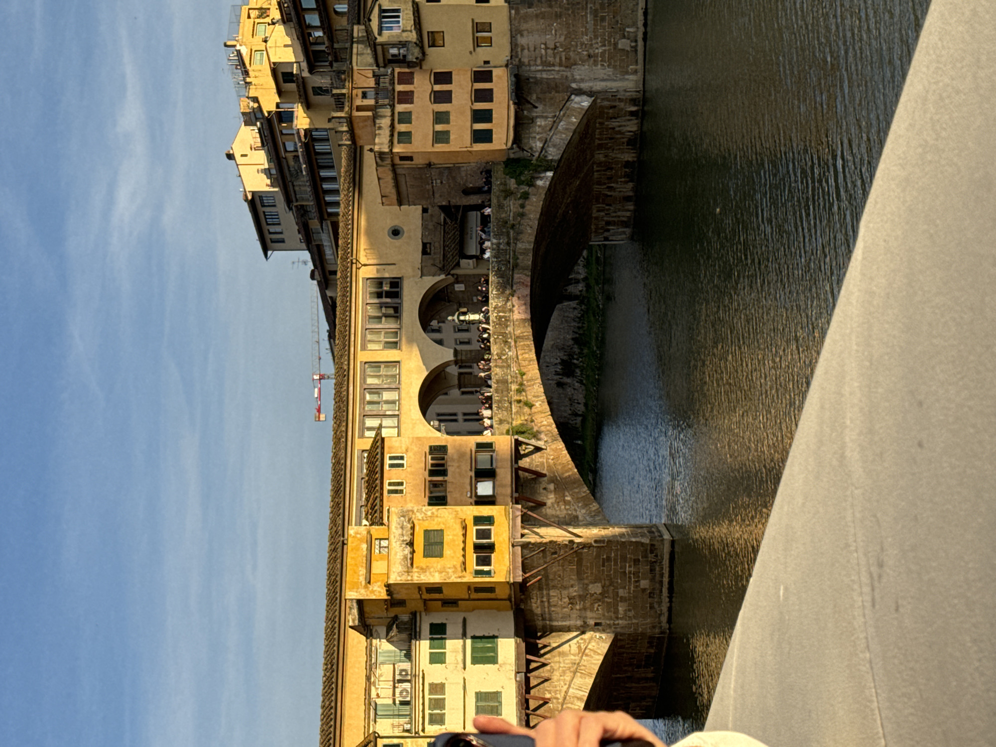 This is a view of Ponte Vecchio in Florence, Italy. The bridge spans a body of water, reflecting the surrounding buildings. The buildings on the bridge are a series of close-set structures, several stories high. Arched openings are visible, along with windows and detailing on the building façades. The sky is blue with sparse clouds. A portion of a bridge and person are in the foreground.