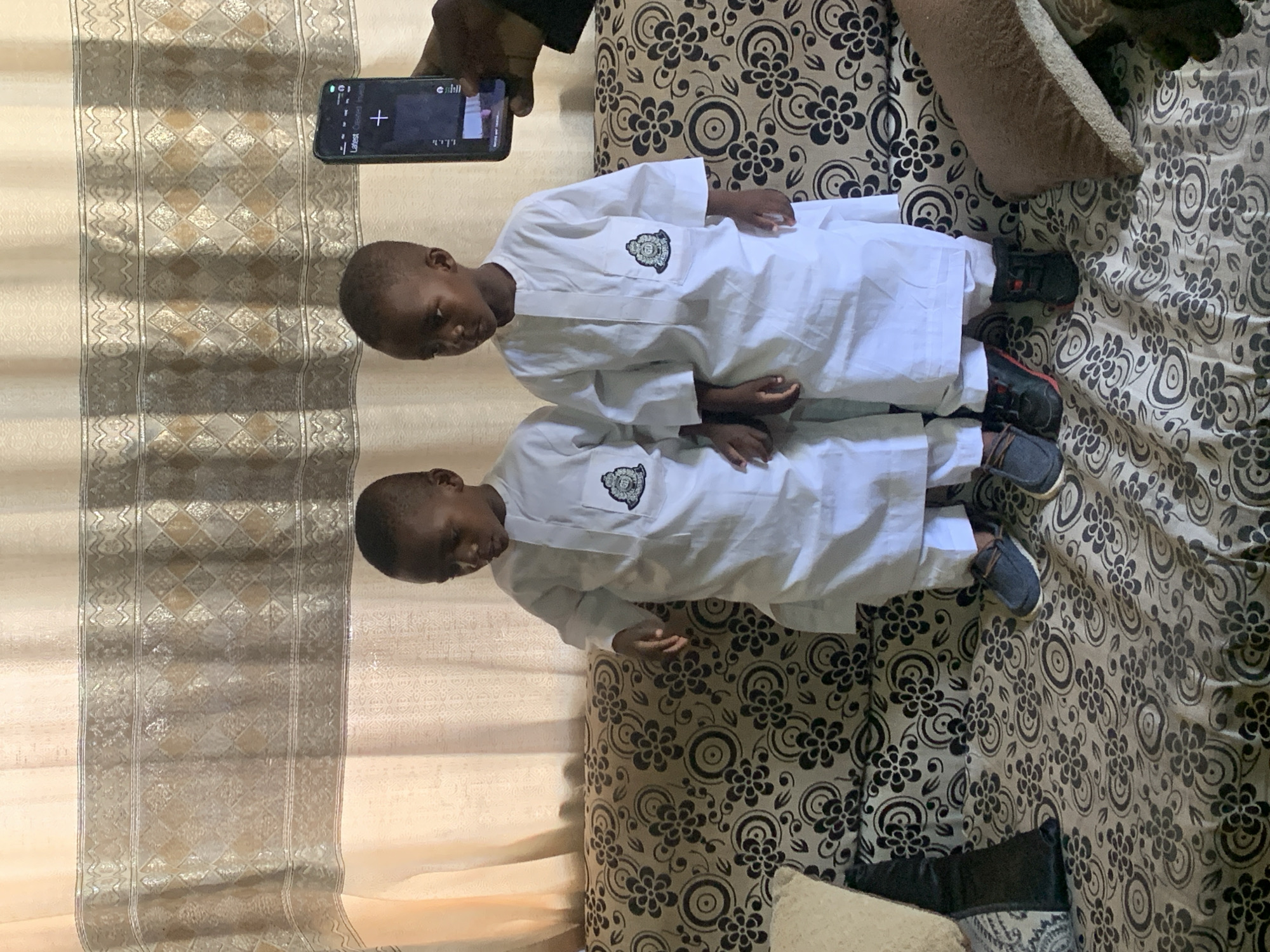 Two young children wearing white shirts with badges are lying side by side on a patterned bedspread, with a phone held above them, presumably to take a picture. The location appears to be in a domestic setting, potentially in Gura Top, Nigeria.