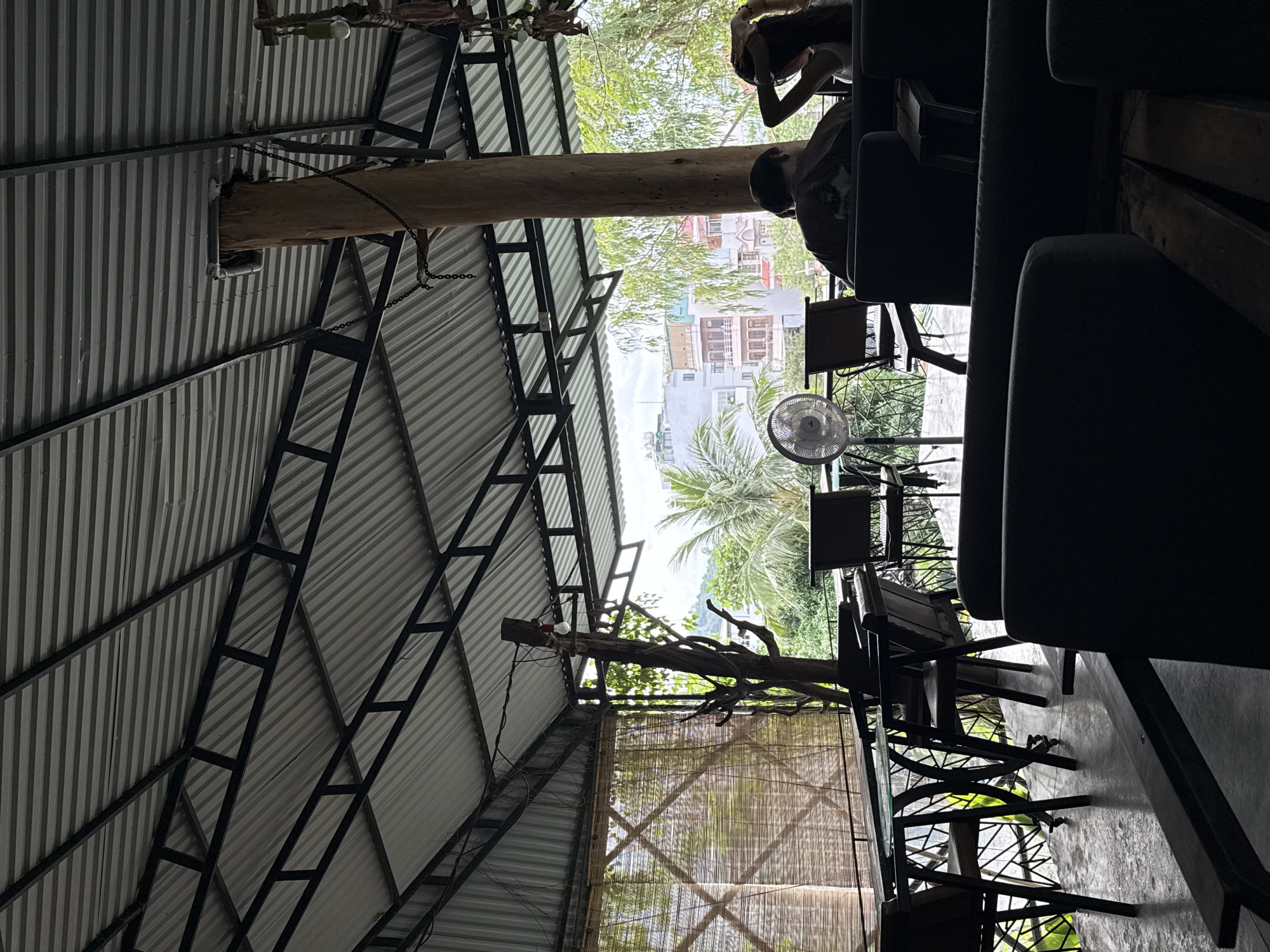 This image, when viewed upright, depicts an open-air cafe or restaurant, likely in Bac Nha Trang Ward, Vietnam. The scene is captured from within the establishment, looking out towards the distant environment.
The interior features a high, sloped ceiling constructed from corrugated metal sheeting supported by a black, angular metal truss framework. A large, natural tree trunk stands prominently within the space, acting as a central pillar or decorative element, with chains and wiring attached.
In the foreground and midground, several dark, upholstered chairs or sofas are arranged with tables, indicating a seating area. Two people are partially visible; one in a dark t-shirt is seen from the back/side, and another with lighter hair is visible leaning forward. A white standing fan is positioned near the tables, suggesting warm temperatures.
Beyond the covered area, the outdoor environment is bright and clear. Lush green trees, including identifiable palm fronds, are prominent. In the background, several multi-story buildings with varied architectural styles line the horizon under a bright sky. A bamboo screen or blind adds to the natural decor on one side.
The time of day appears to be sunny daytime, likely mid-morning or afternoon, given the bright outdoor light and visible shadows. The overall setting conveys a relaxed, casual atmosphere. No text is visible in the image.