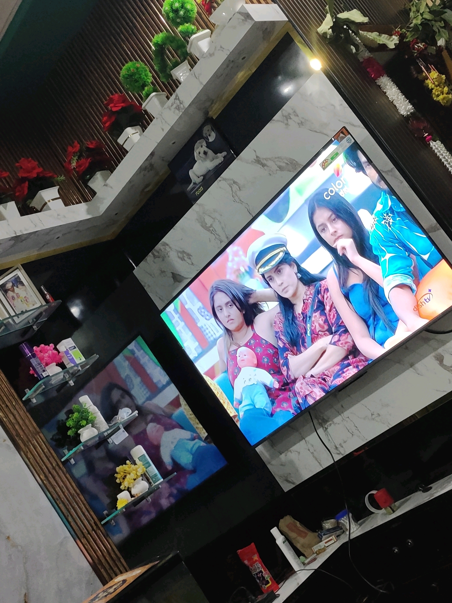 The image shows an indoor scene, possibly a living room or entertainment area, with a large television screen displaying a scene from a television program featuring three young women. The television is mounted on a wall with a marble-like finish. Below the television are several shelves with decorative items and toiletries. In the background, there are decorative plants and what appear to be garlands or festive decorations. The overall lighting suggests it's daytime or evening with artificial lighting. The composition is angled, giving a dynamic perspective of the room.