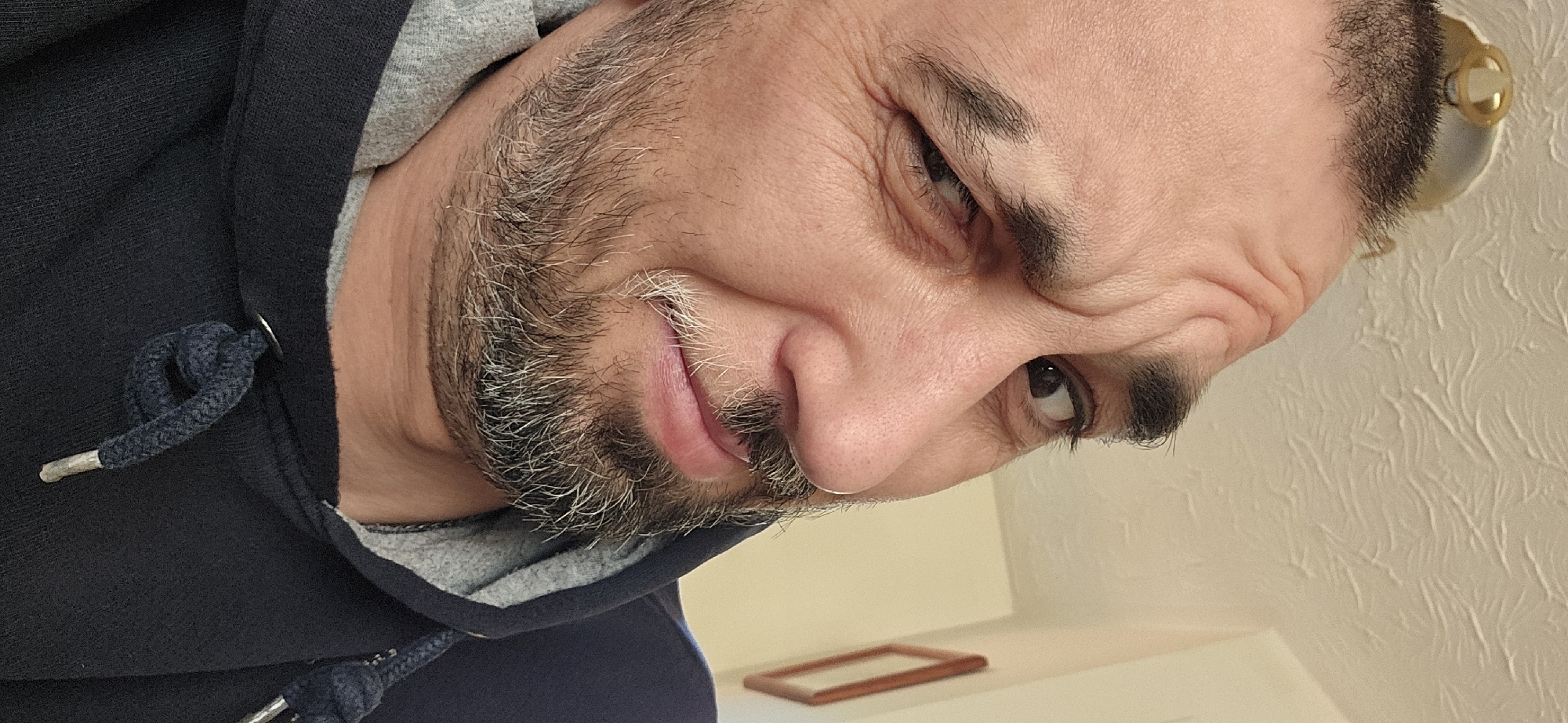 This close-up portrait, rotated approximately 45 degrees counter-clockwise, features an adult male with light-to-medium skin tone. He has a dark beard and mustache with prominent grey and white hairs, especially around his mouth. His dark eyebrows are visible, and his right eye (viewer's left) is closed in a distinct wink or squint, creating noticeable wrinkles around it, while his left eye is open and looking upwards. A subtle, playful smile is hinted at the corner of his mouth, giving him a mischievous or humorous expression.
He is wearing a dark blue or black hooded sweatshirt (hoodie) with a grey inner lining visible near his neck. A dark blue drawstring with silver-toned metal tips and a silver eyelet is prominently featured on his clothing.
The background consists primarily of a light-colored wall or ceiling with a distinct textured pattern, suggesting anaglypta wallpaper or a similar decorative finish. In the lower right section of the image, a small, empty rectangular wooden frame rests on a light surface, possibly a shelf. A glimpse of a gold-colored light fixture or decorative element is visible in the upper right. The scene is set indoors, likely within a home environment, consistent with a location such as Norwich, United Kingdom. There is no visible text.