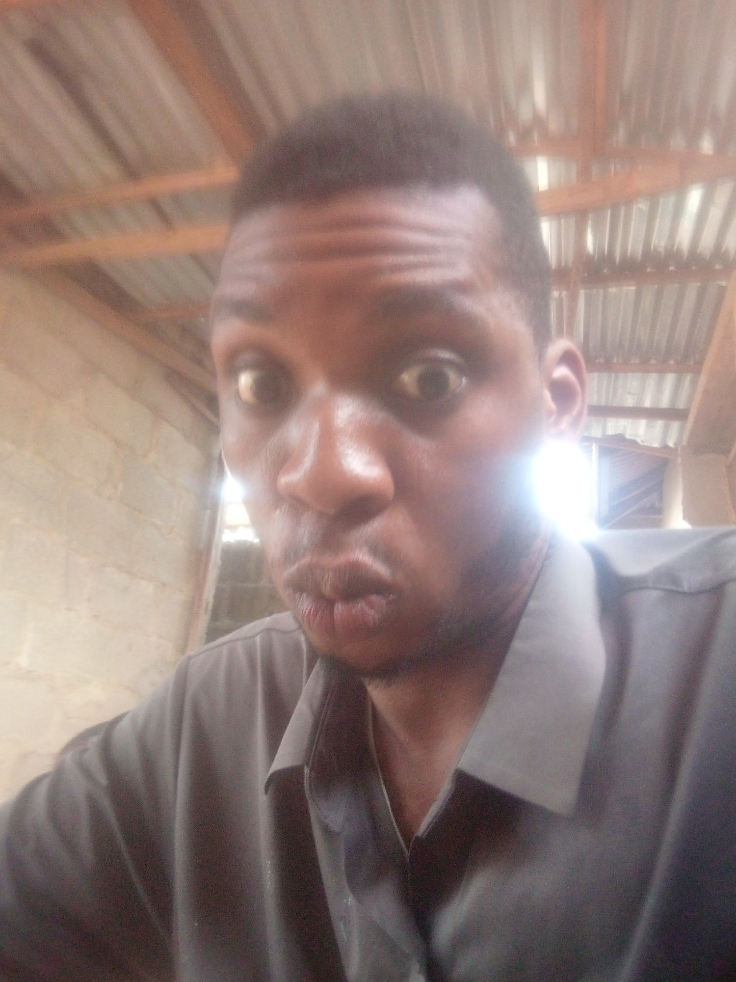 A close-up, eye-level shot captures a man of dark complexion making a humorous or surprised face. He has wide-open eyes and pursed lips, often referred to as a "duck face." His short, dark hair and some stubble on his chin are visible. He is wearing a dark grey or black collared shirt.
The man dominates the foreground, with his face angled slightly towards the camera. The background suggests an indoor or semi-enclosed space. Above his head, a light-colored corrugated metal roof with visible wooden support beams can be seen. To his left, a wall constructed from light-colored concrete blocks is visible. A bright light source, likely natural sunlight, is prominent behind and to the right of the man, causing a strong glare and overexposed area in that section of the image. This light illuminates the right side of his face.
The overall lighting is bright, but the image appears somewhat grainy and soft in focus. No text is visible in the image.