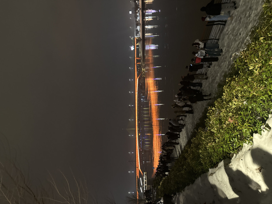 A long bridge illuminated with orange lights spans across a dark body of water, its reflection visible. A crowd of people stand along the edge of the water, seemingly observing the bridge. The right side of the image is lined with lush greenery and a white, possibly snowy, embankment. The sky is dark and overcast, adding to the nighttime atmosphere.