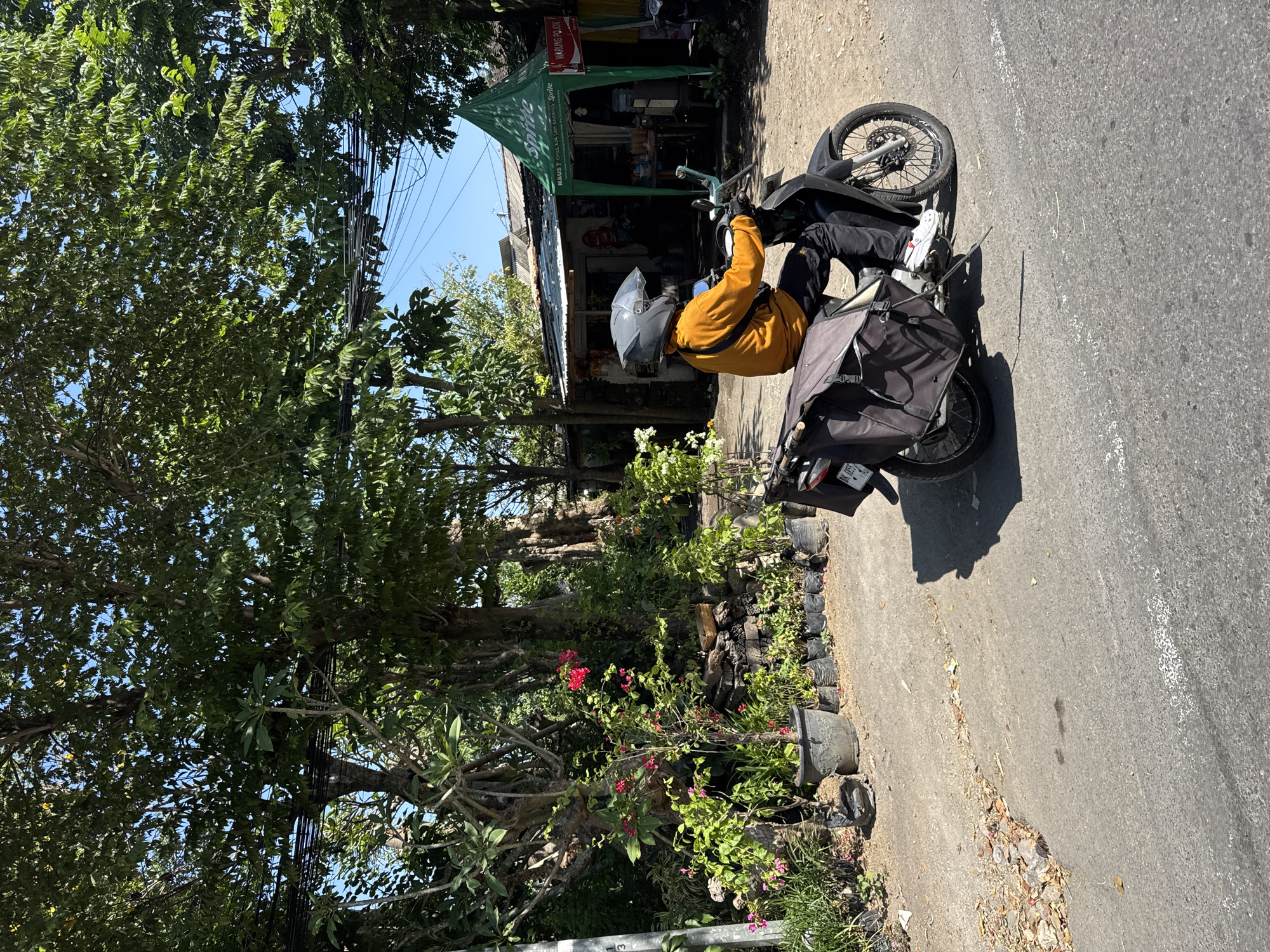 A person wearing a yellow long-sleeved shirt, black pants, and a grey helmet is riding a black motorcycle on a paved road. The motorcycle has a large black bag attached to the rear. The person is wearing white sneakers. The scene appears to be in Denpasar, Indonesia, as indicated by the presence of tropical foliage, a small shop with a green tent bearing the "Sprite" logo, and a license plate that starts with "DK," a common prefix for vehicles registered in Bali, Indonesia. The shop appears to be a small local business, possibly a warung or convenience store, with goods visible on display. The environment is outdoors, under a clear blue sky, with trees and plants lining the roadside. The road surface shows signs of wear and tear, with some debris scattered on it. The overall impression is of a typical street scene in a warm, possibly tropical, urban or semi-urban setting.