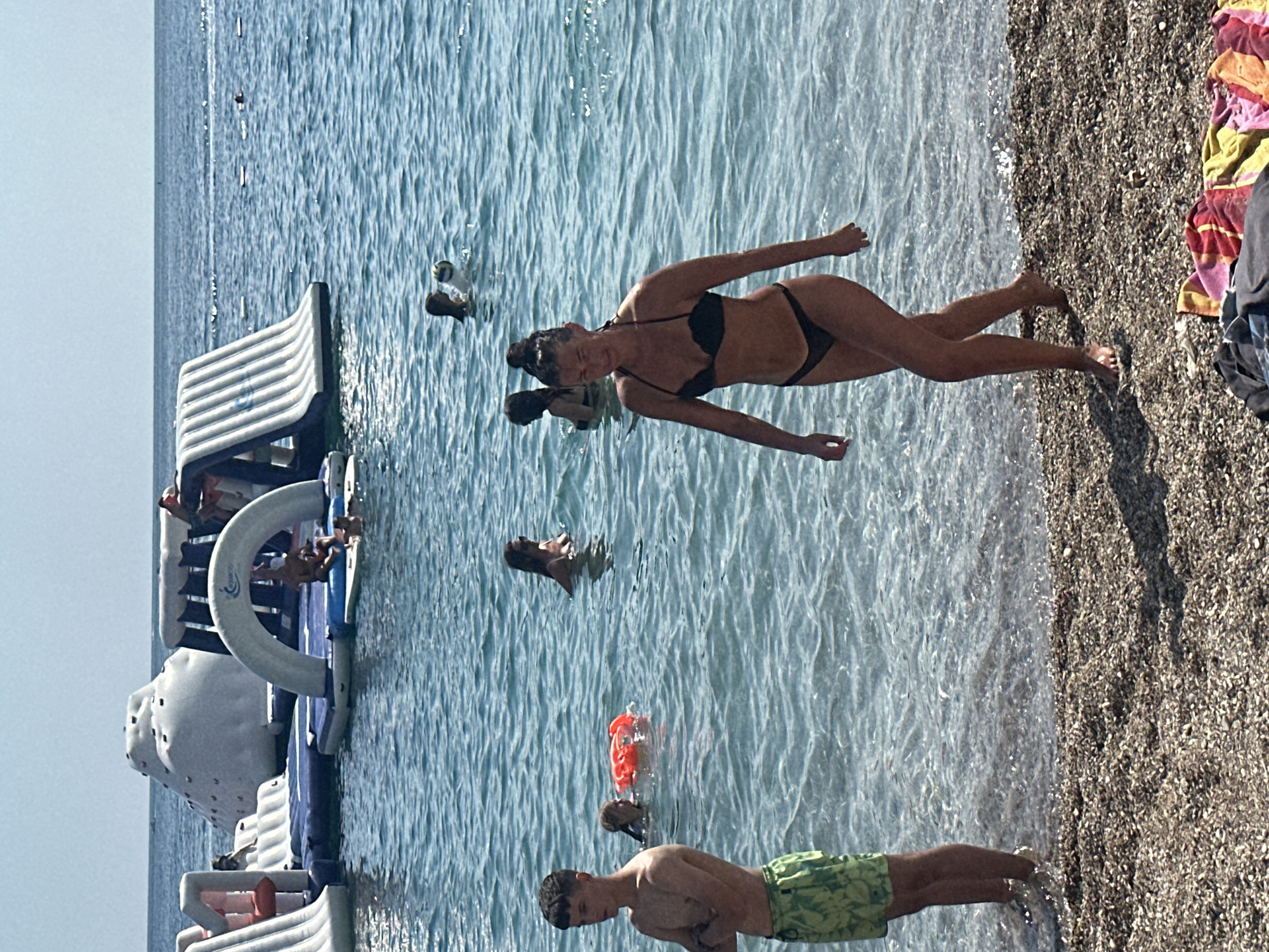 In the city of Budva, Montenegro, on a sunny day at the beach, a young man stands on the shore wearing green patterned shorts. Further into the clear blue water, a woman in a black bikini walks towards the beach, her body angled away from the viewer. In the middle ground, several people are enjoying the water, including a woman with a red inflatable ring, a woman wearing a black bikini, and a group of people near a large inflatable water park in the background. The water park features slides and climbing structures, with more people visible enjoying the attractions. The beach is covered in pebbles, and towels are laid out on the sand in the foreground. The sea stretches out to the horizon under a clear sky.
