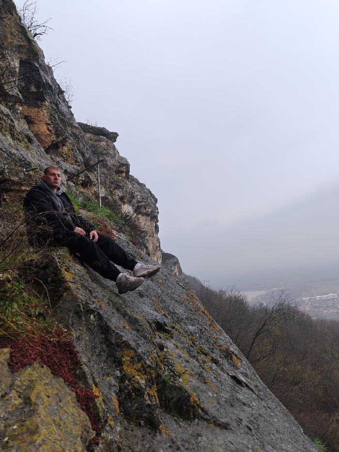 A male individual, approximately 20-30 years of age, is seated on a steep, weathered limestone cliff face on the Shumen Plateau, overlooking the city of Shumen, Bulgaria. He wears a dark jacket, dark trousers, and light-grey athletic shoes, with a grey scarf visible around his neck. His posture is relaxed, with legs extended downwards along the rock slope, and his gaze directed towards the left. The rock face exhibits varying textures with patches of green and yellow lichen, and areas of reddish ground cover. Two vertical metal poles are partially visible embedded in the rock behind the subject. The background presents an expansive view of a valley containing urban development under an overcast grey sky. Bare deciduous trees are present on the slopes below the subject.