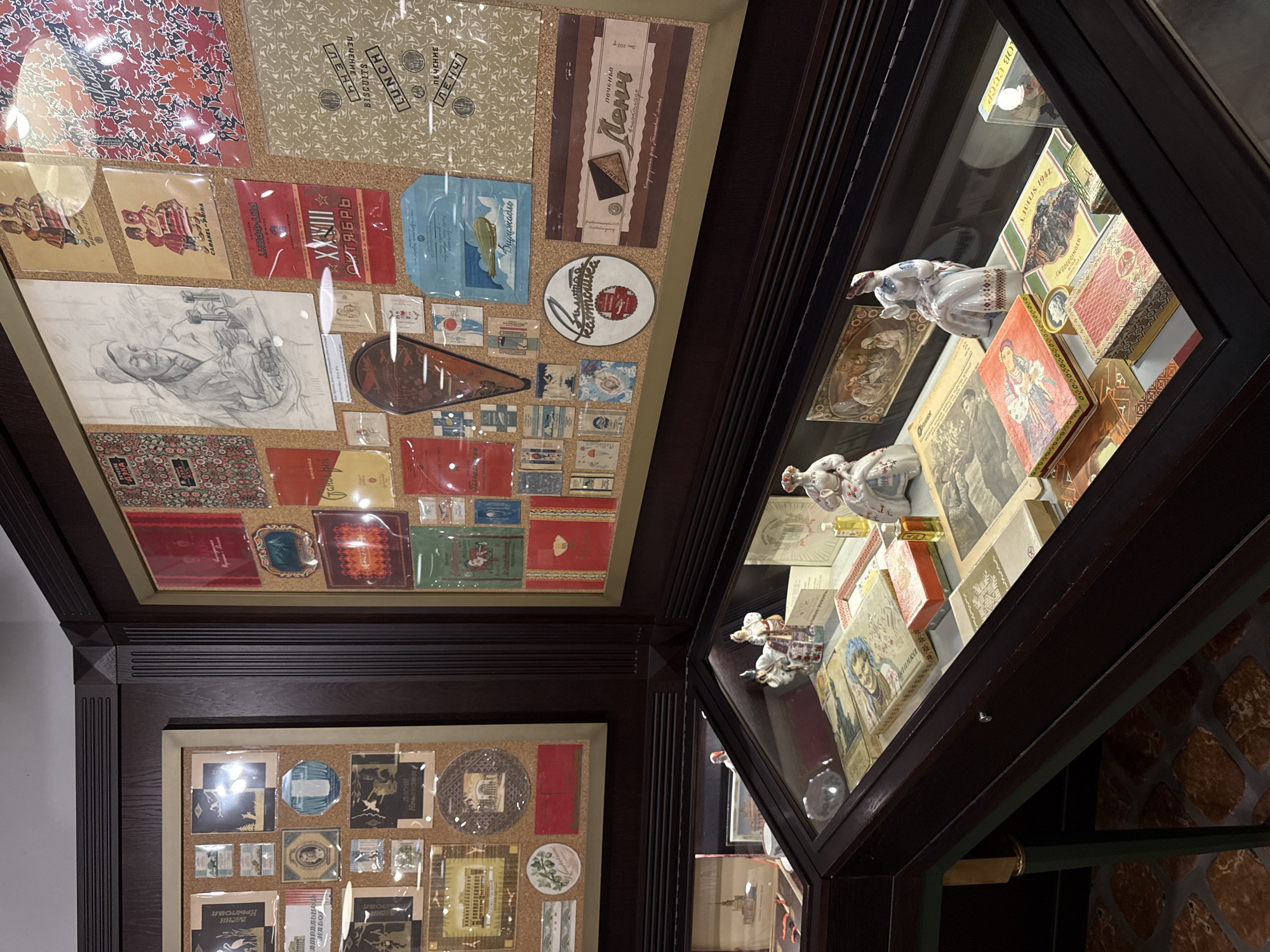 The image captures a display case within a museum in Arkhangel'skoe, Russia. The display case is filled with antique boxes, packaging, and small figurines. Among the items are boxes with intricate designs featuring people in traditional clothing, as well as packaging for what appears to be candy or biscuits. There are also small ceramic figurines of women in traditional attire. The display case itself is made of dark wood, with a glass top. In the background, a large wall display features a collection of vintage posters, advertisements, and artwork, also mounted on a corkboard-like material. The overall environment suggests a historical exhibition, likely showcasing Soviet-era or earlier Russian consumer goods and art.