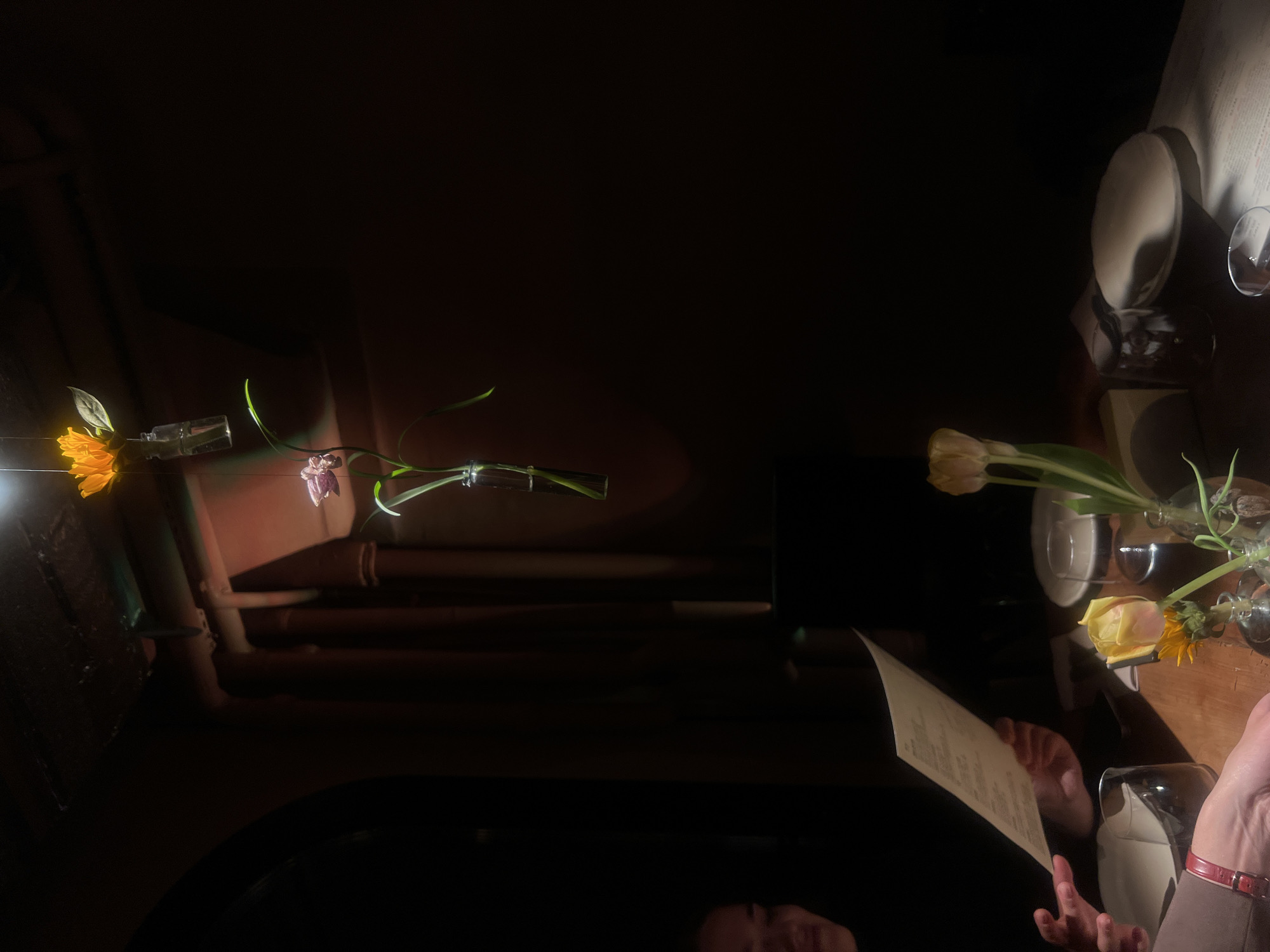 The image shows a dimly lit restaurant in Moscow, Russia. A table is set with plates, wine glasses, and small vases containing flowers. A person is holding a menu, seemingly contemplating their order. The table reflects the lighting and some of the surrounding environment. The background is dark, but hints of architectural features can be seen.