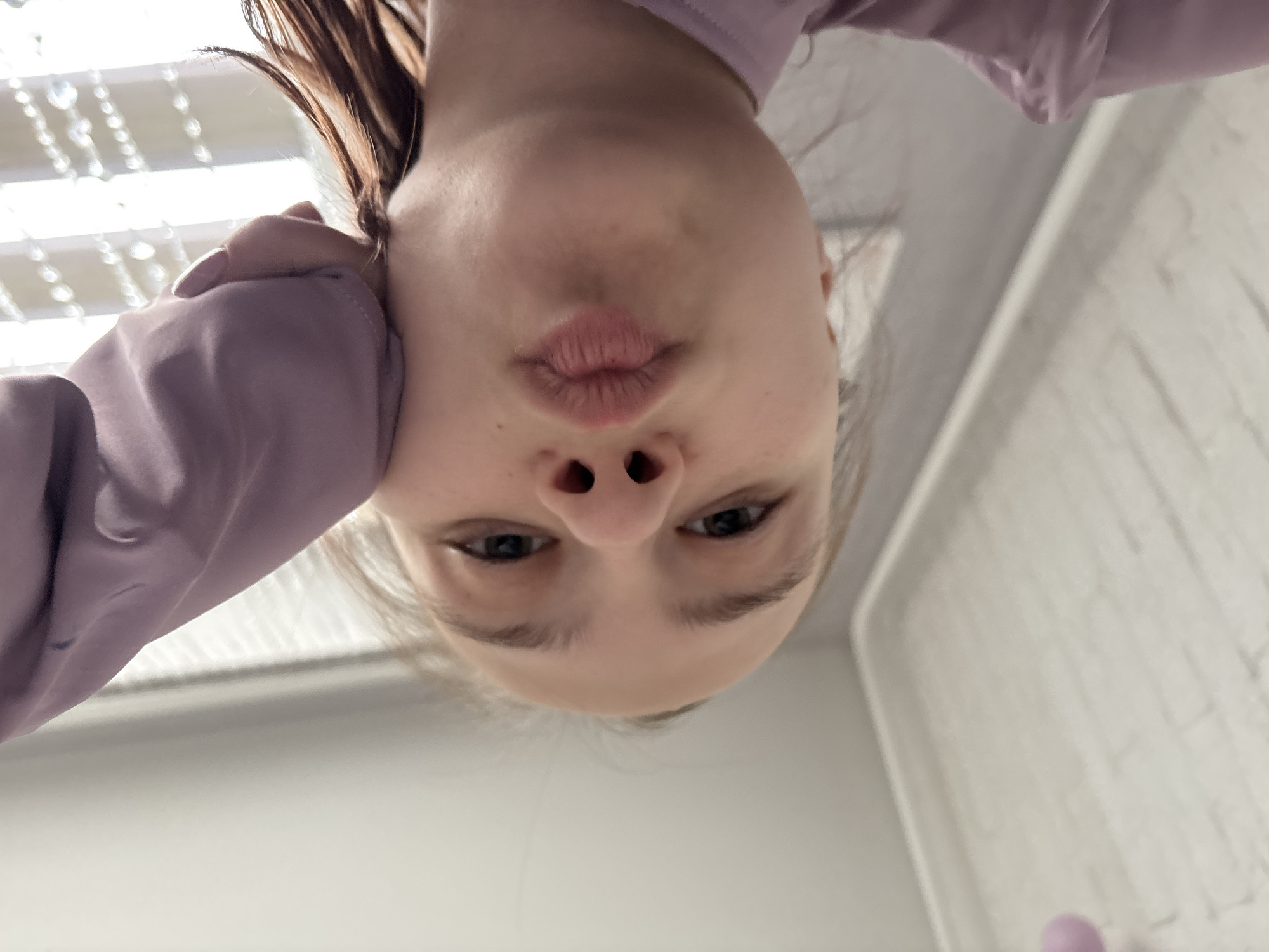 A young woman is captured in a close-up, downward-angled selfie. Her lips are pursed, suggesting a kiss. She has fair skin, dark eyes, and light brown hair visible at the top left. She is wearing a light purple, long-sleeved garment. The background includes a blurred beaded curtain on the left and a textured white wall on the right. The location is not discernable. There is no information about the city of Chaikovsky, Russia.